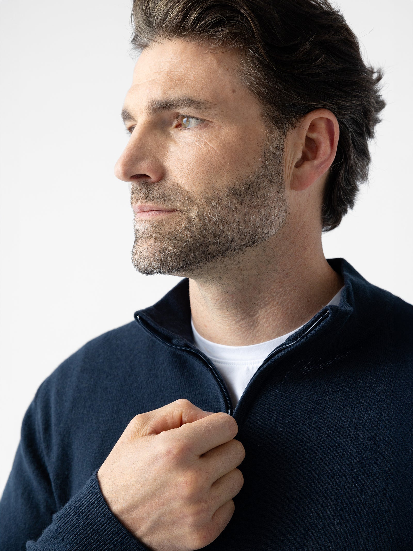 A man with short, dark hair and a beard gazes thoughtfully into the distance. He is dressed in a Cozy Earth Men's Quarter Zip Sweater, which he is partially unzipping with one hand. The plain white background emphasizes his contemplative expression. 