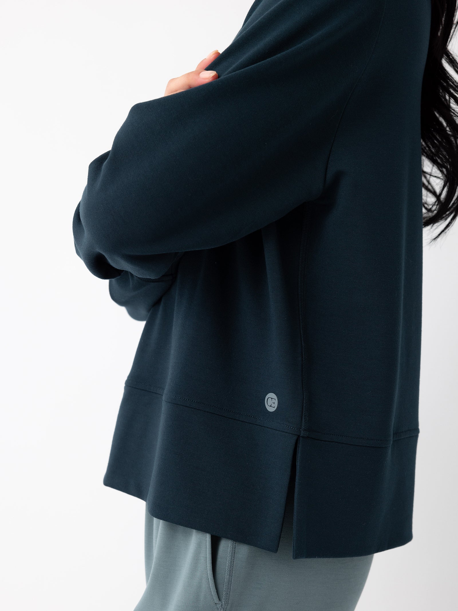 Close-up of a person wearing a Cozy Earth Women's StudioLite Crewneck in navy blue and grey pants. The person is standing sideways with their arms crossed, displaying only the upper half of their body against a plain white background. The Crewneck features a small logo near the hem. 