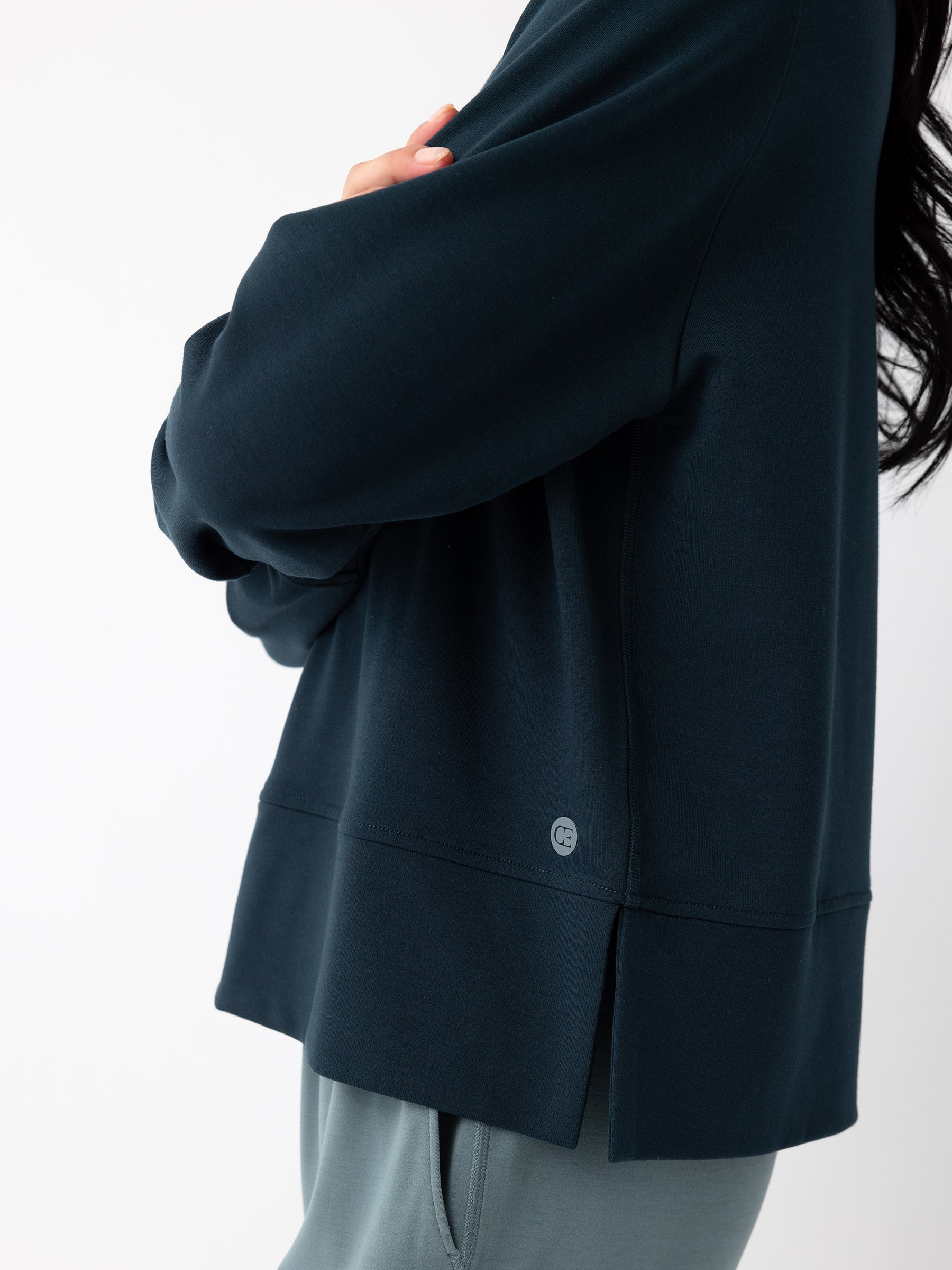 Close-up of a person wearing a Cozy Earth Women's StudioLite Crewneck in navy blue and grey pants. The person is standing sideways with their arms crossed, displaying only the upper half of their body against a plain white background. The Crewneck features a small logo near the hem. |Color:Eclipse