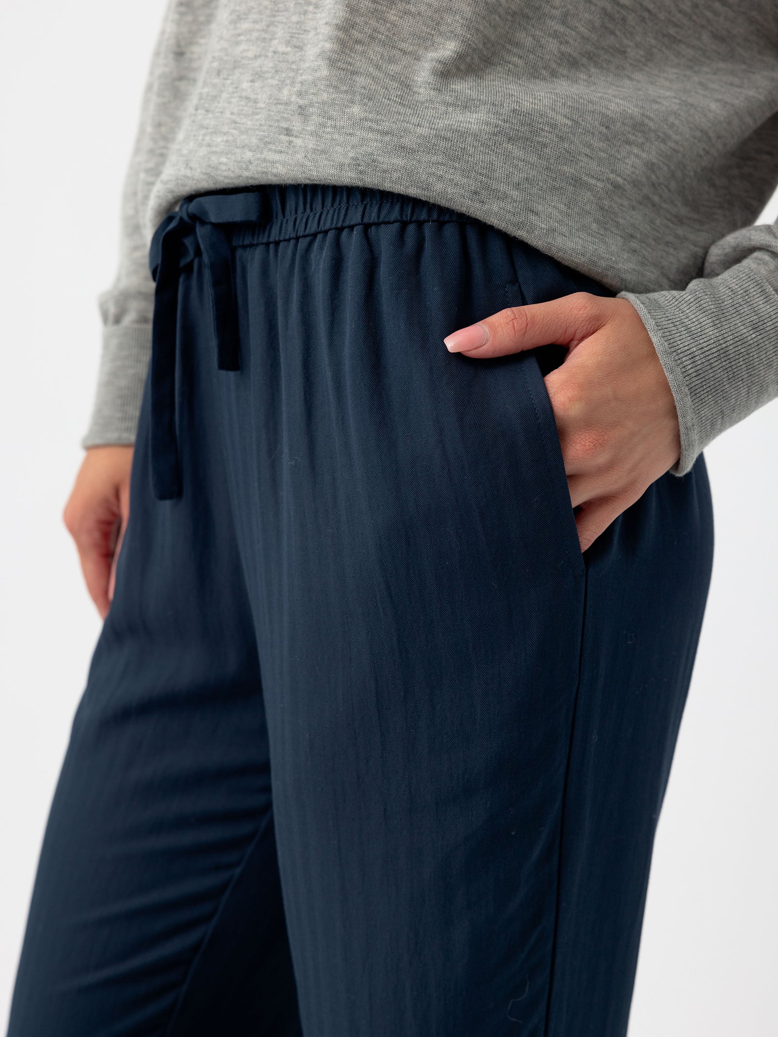 A person wearing a grey sweater and Cozy Earth's Women's Sunset Cropped Pant in dark blue, featuring a loose fit and an elastic waistband, stands with one hand in their pocket. The image highlights the midsection and part of the arm, showcasing the relaxed fit and texture of the clothing. 