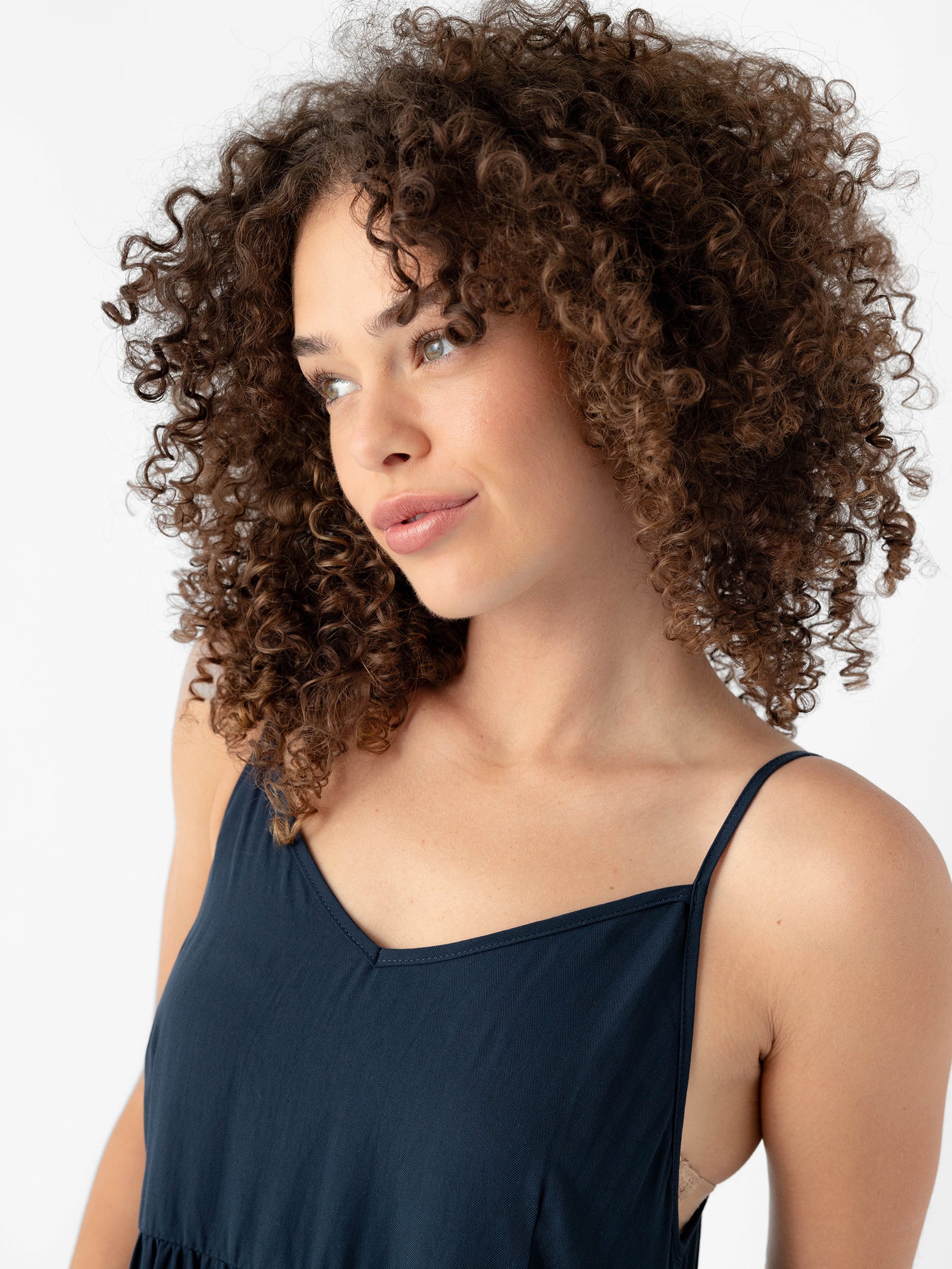 A person with curly hair wearing a navy blue Women's Sunset Gathered Dress from Cozy Earth poses against a white background. The individual looks slightly to the side with a calm expression on their face. 