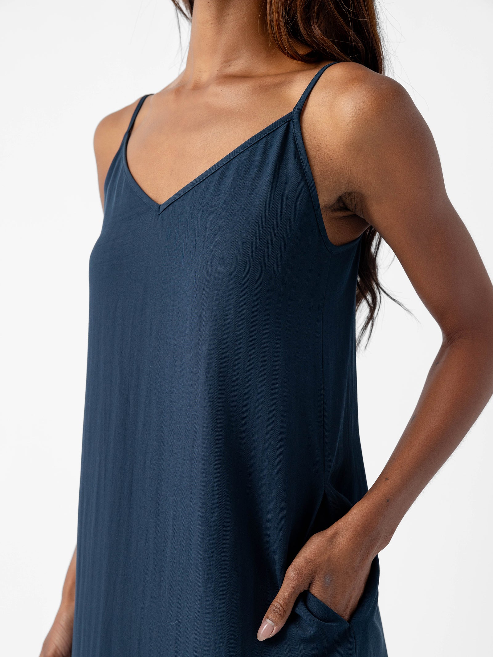 A person with long hair is wearing the Women's Sunset Slip Dress from Cozy Earth, which is a dark blue, sleeveless dress featuring thin shoulder straps and a V-neckline. The person has one hand in their pocket, standing against a plain white background. 