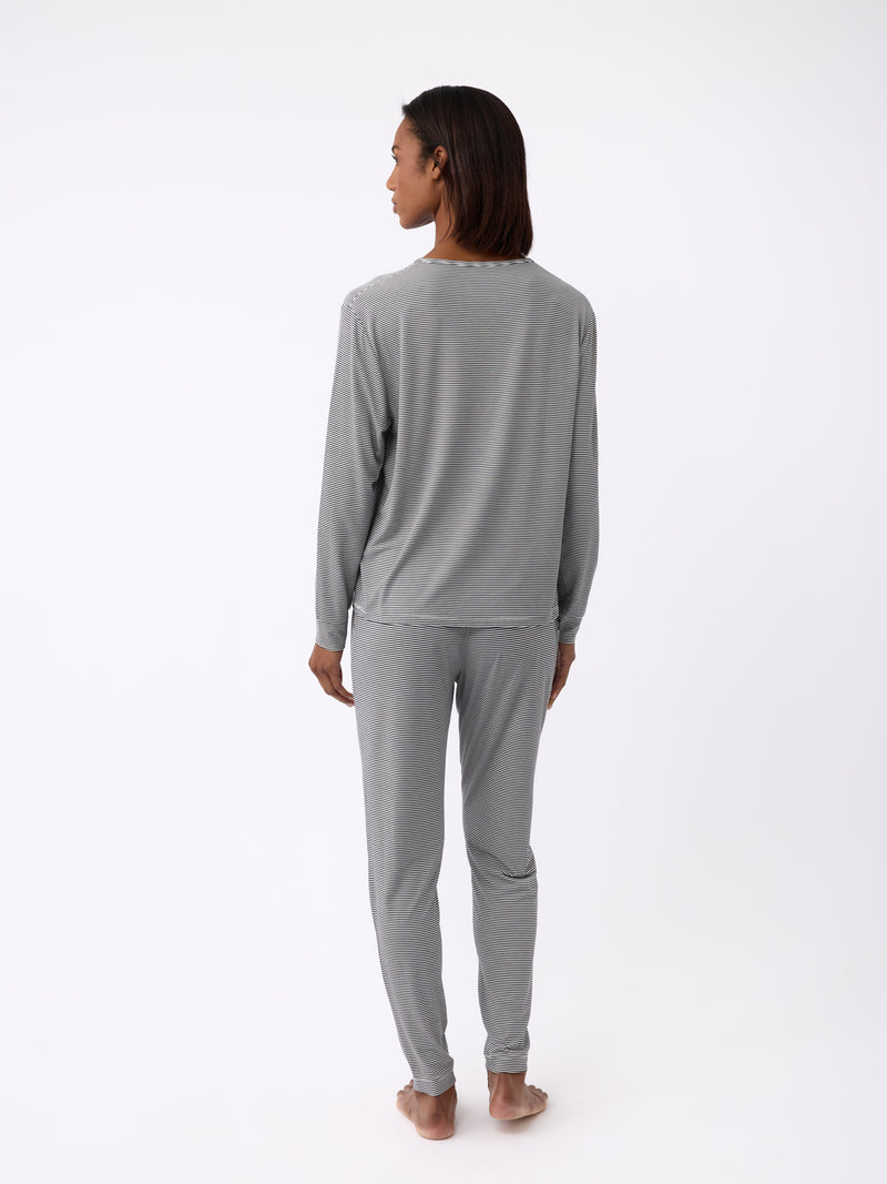 A person stands barefoot on a plain white background, wearing the Cozy Earth Women's Bamboo Stretch-Knit Long Sleeve Lounge Tee in light gray pajamas. They're facing away to show the back of the soft and comfortable set.