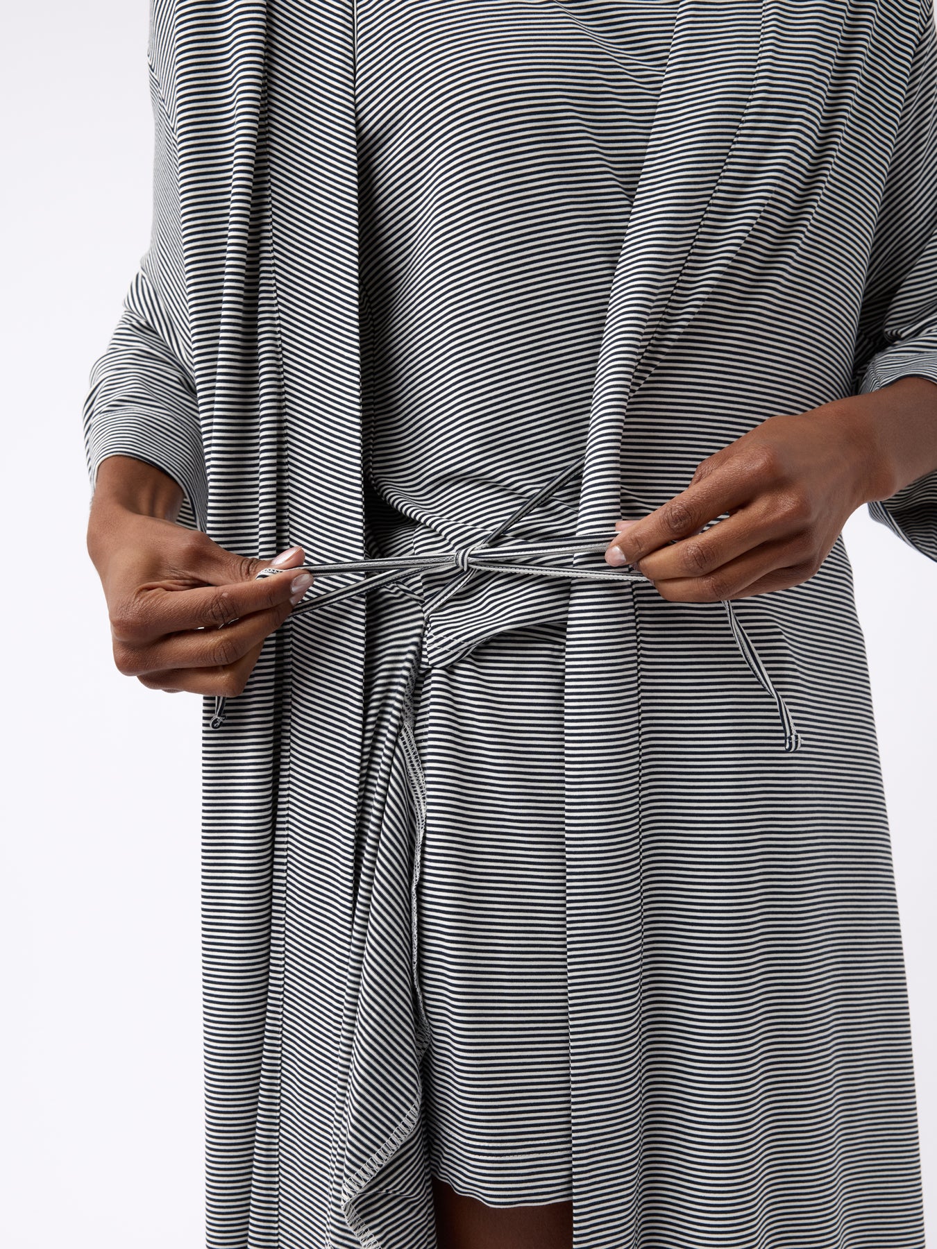 A person fastens the belt of a Cozy Earth Women's Bamboo Stretch-Knit Kimono Robe in black and white stripes. The lightweight robe has long sleeves, and the setting features a plain, light-colored background. 