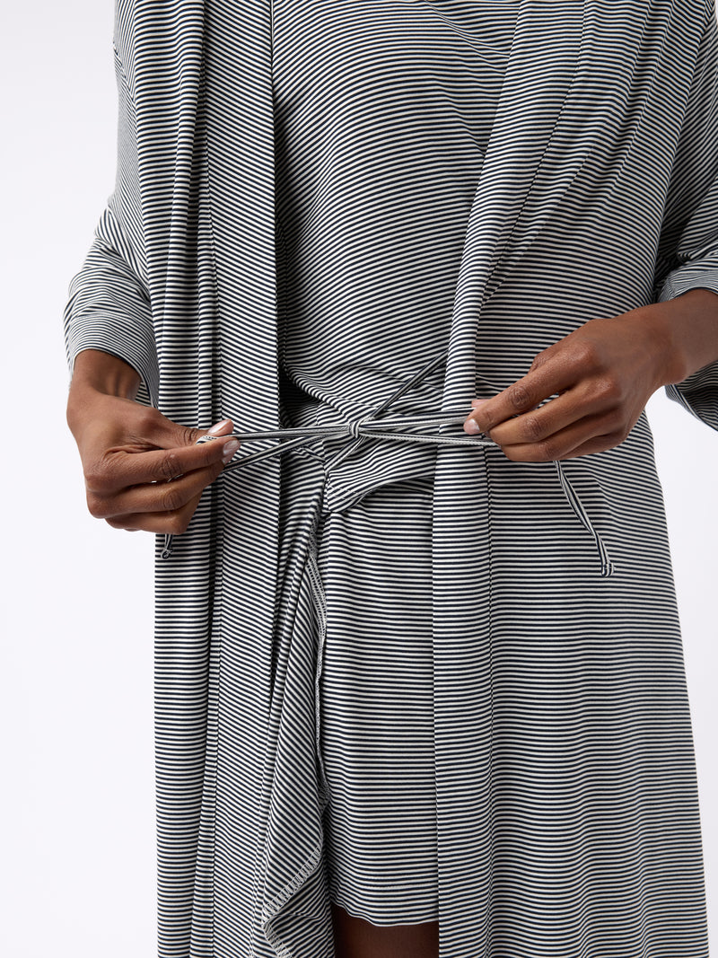 A person fastens the belt of a Cozy Earth Women's Bamboo Stretch-Knit Kimono Robe in black and white stripes. The lightweight robe has long sleeves, and the setting features a plain, light-colored background.