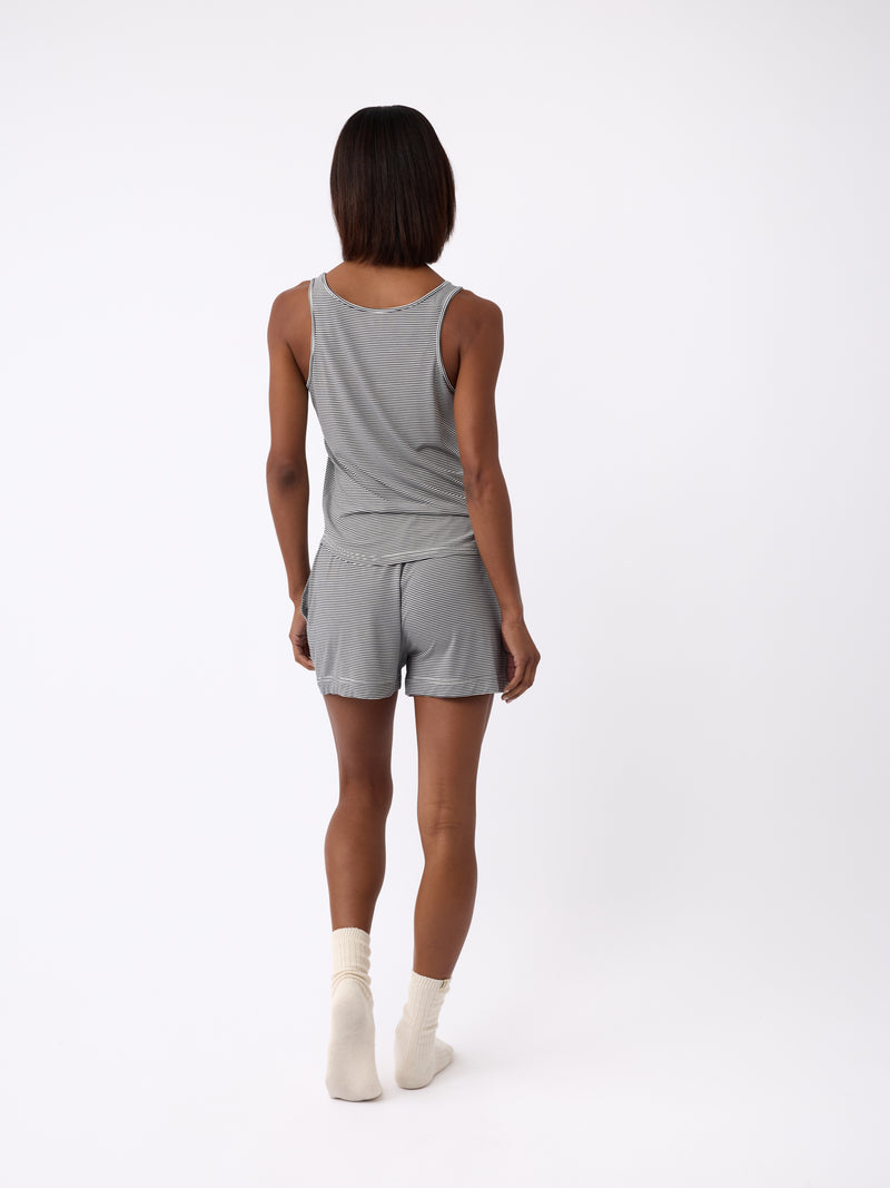 A person faces away, clad in a gray tank top, Cozy Earth's Women's Bamboo Stretch-Knit Shorts, and white socks. They sport short, straight hair against a plain backdrop.