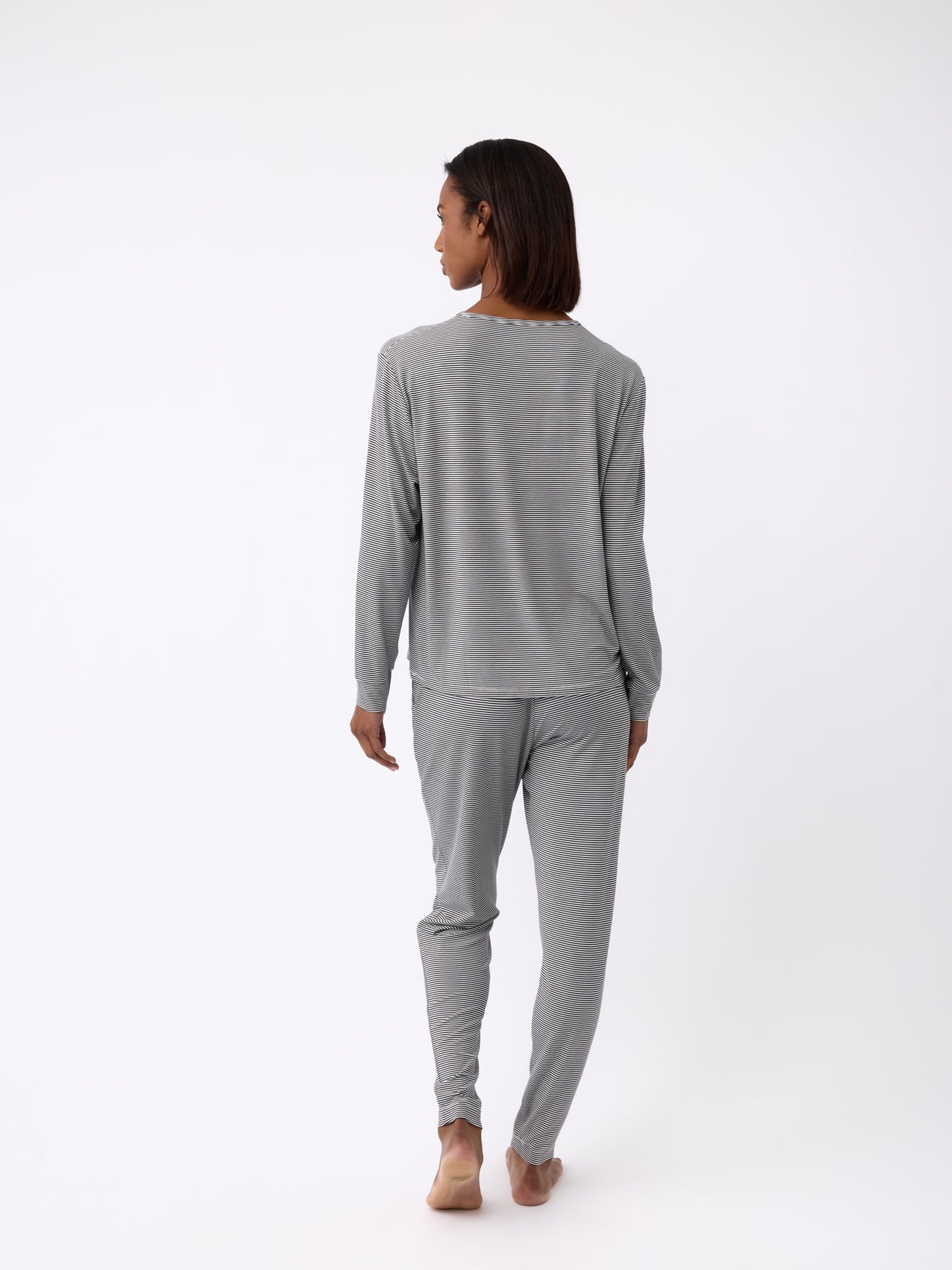 A person stands barefoot, facing away, wearing Cozy Earth's Women's Bamboo Stretch-Knit Tapered Pant in gray loungewear on a white background. The outfit includes a long-sleeve top and pants. 