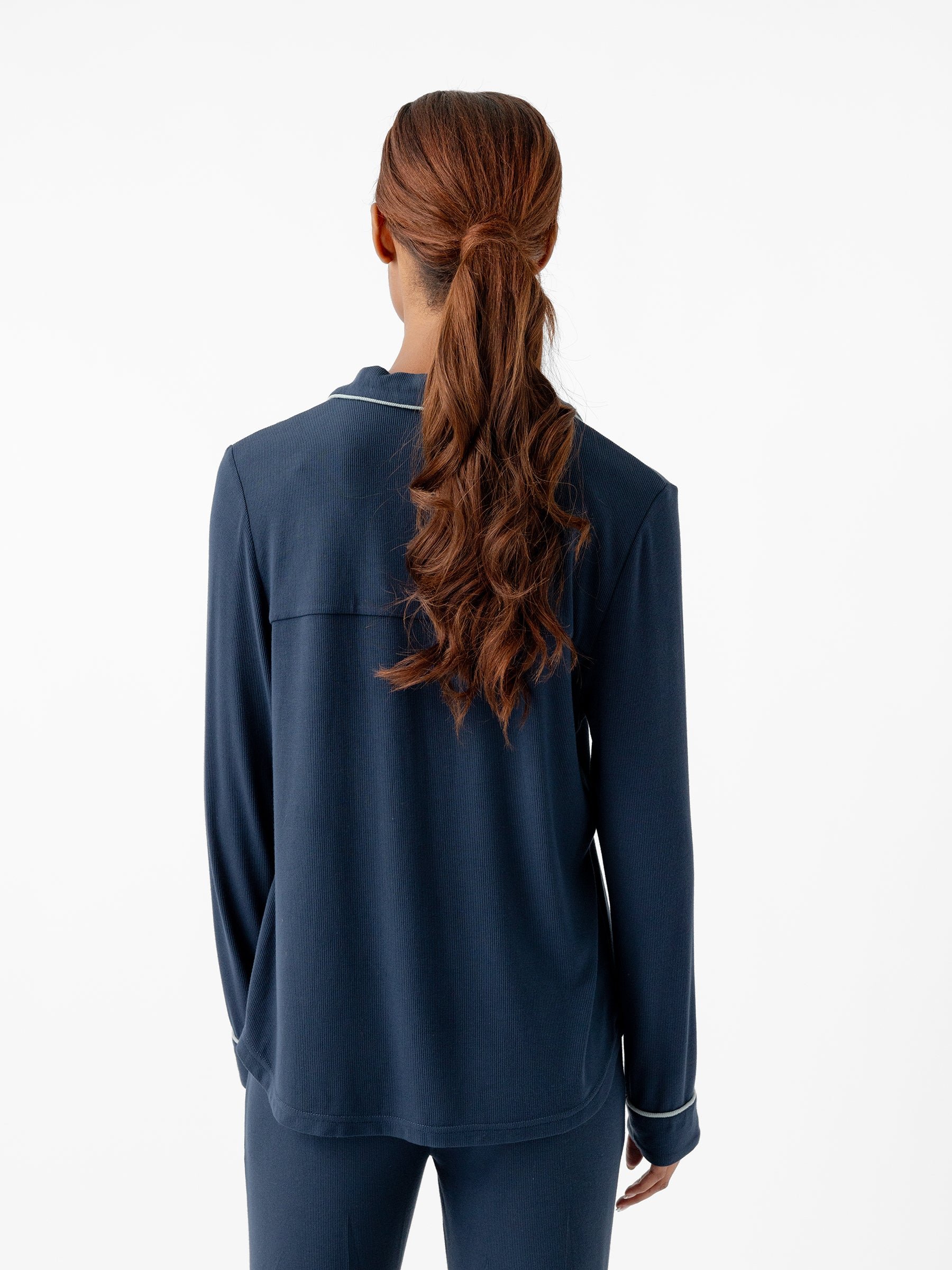 A person with long, wavy brown hair tied in a ponytail is shown from the back. They are wearing a Women’s Bamboo Rib Knit Classic Long Sleeve Pajama Top and matching pants in dark blue, both by Cozy Earth. The background is plain white. 