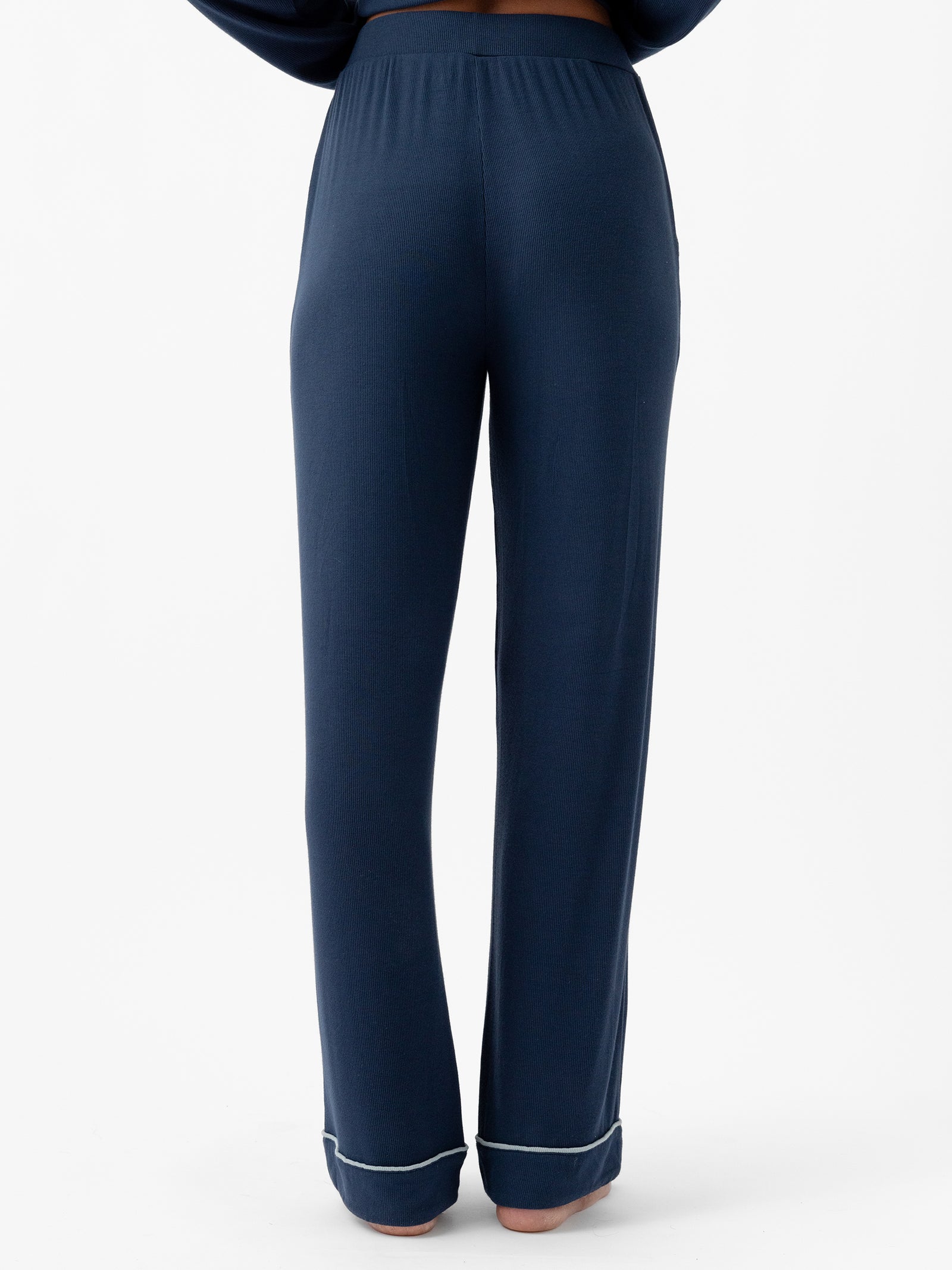 A person is standing with their back to the camera, wearing the Women's Bamboo Rib Knit Classic Pajama Pant from Cozy Earth in navy blue. The full-length pants have a slightly loose fit and feature light-colored piping at the hem. The background is plain white. 