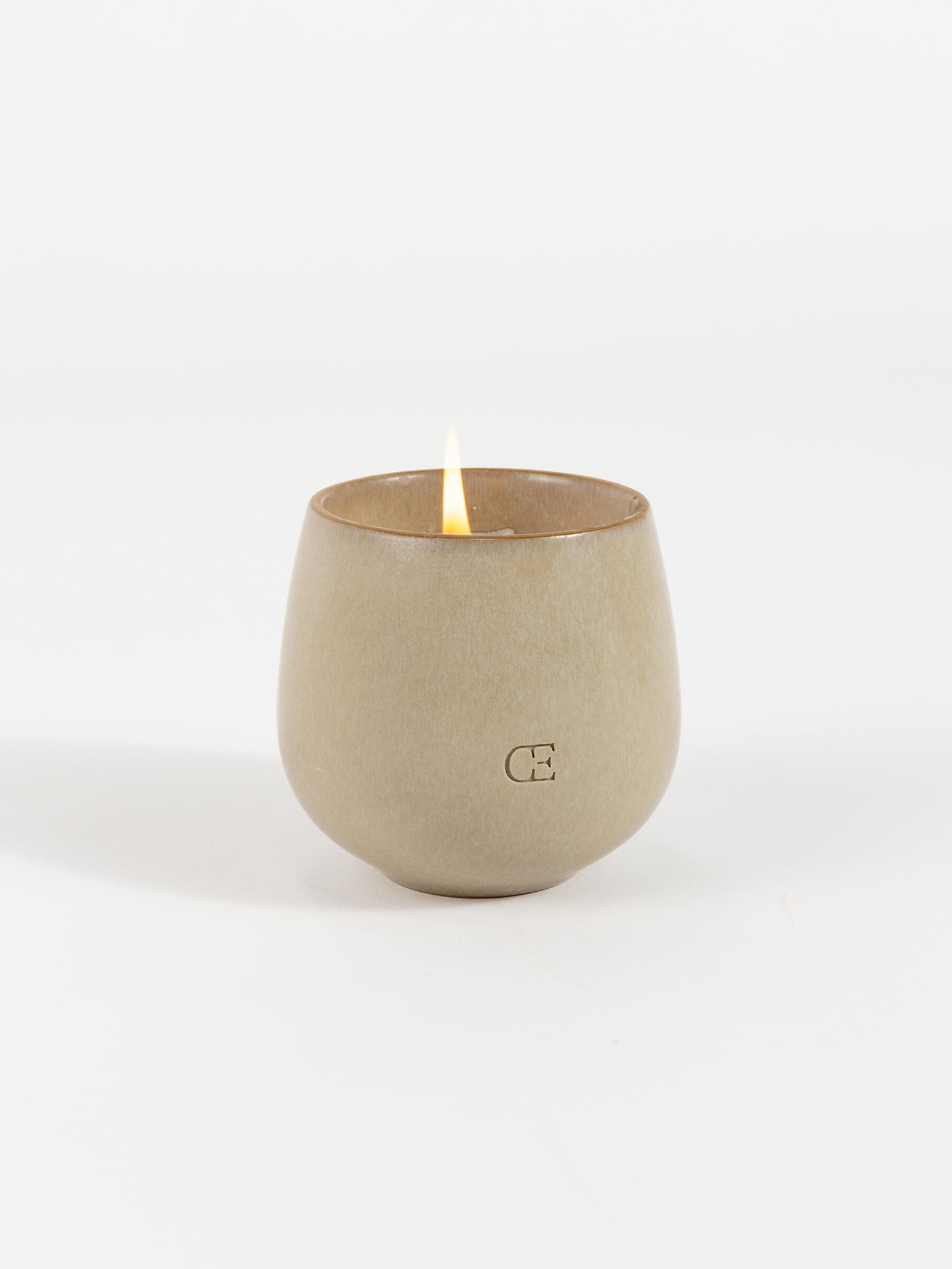The Candle EMBER by Cozy Earth is a beige ceramic holder with a minimalist design, featuring an embossed "C" and a lit candle inside, set against a plain white background.