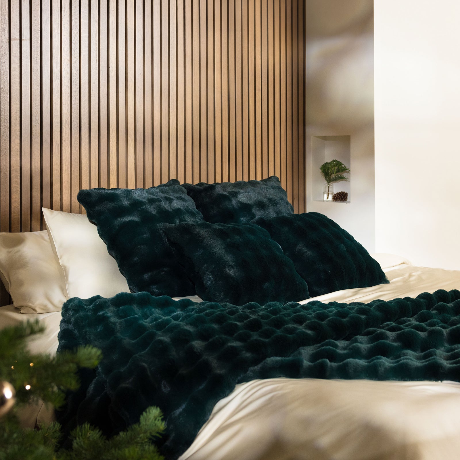 A cozy bedroom featuring a bed adorned with Bubble Cuddle Pillows by Cozy Earth in deep green velvet and a matching textured blanket. The headboard is wooden with vertical slats, and there's a small plant on a white shelf, adding a touch of greenery. 