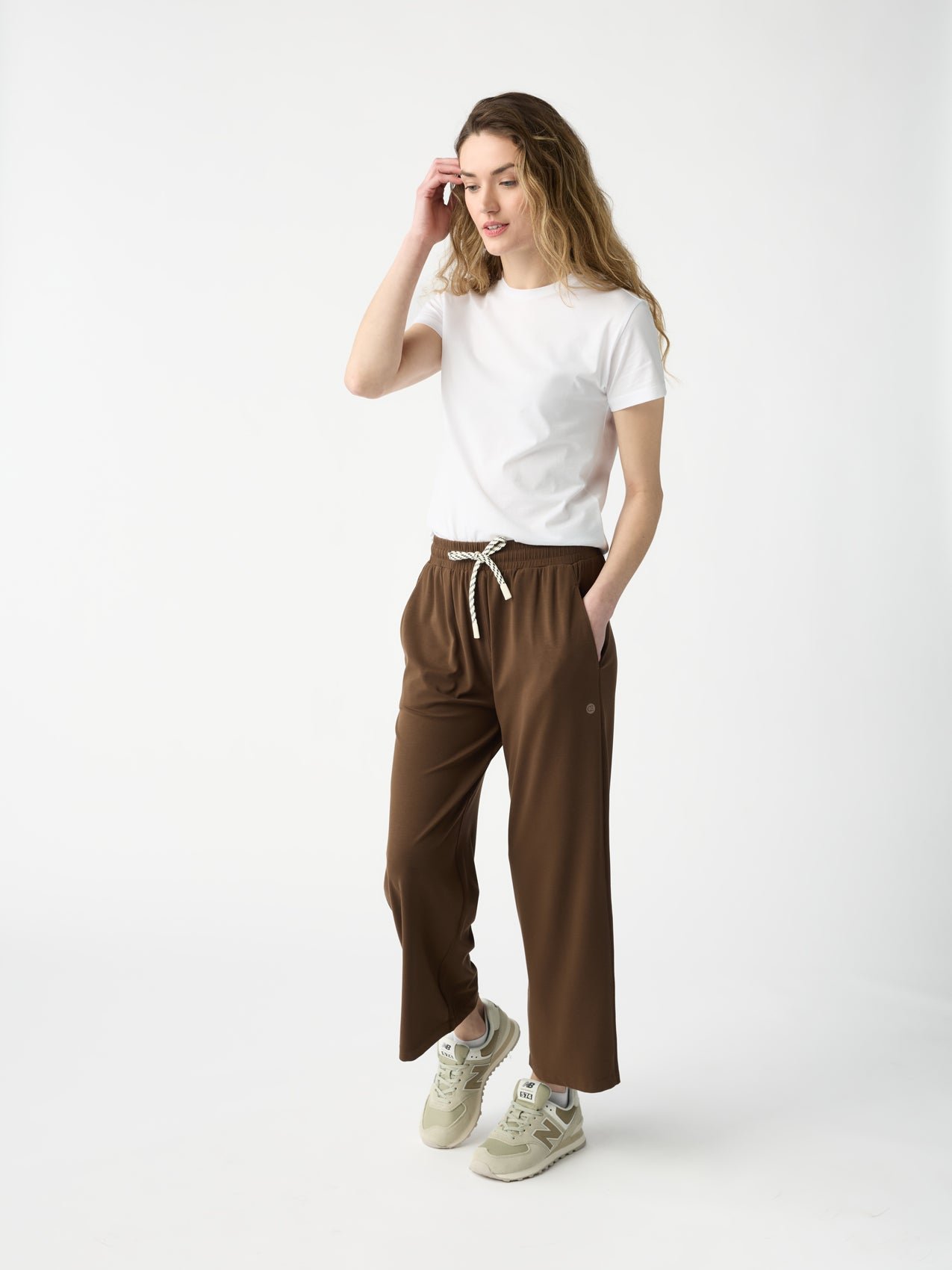 A person with long hair poses against a plain background, wearing a white t-shirt and Cozy Earth Women's Studio Cropped Wide Leg Pants. They sport beige sneakers, one hand in a pocket and the other gently touching their hair. |Color:Espresso