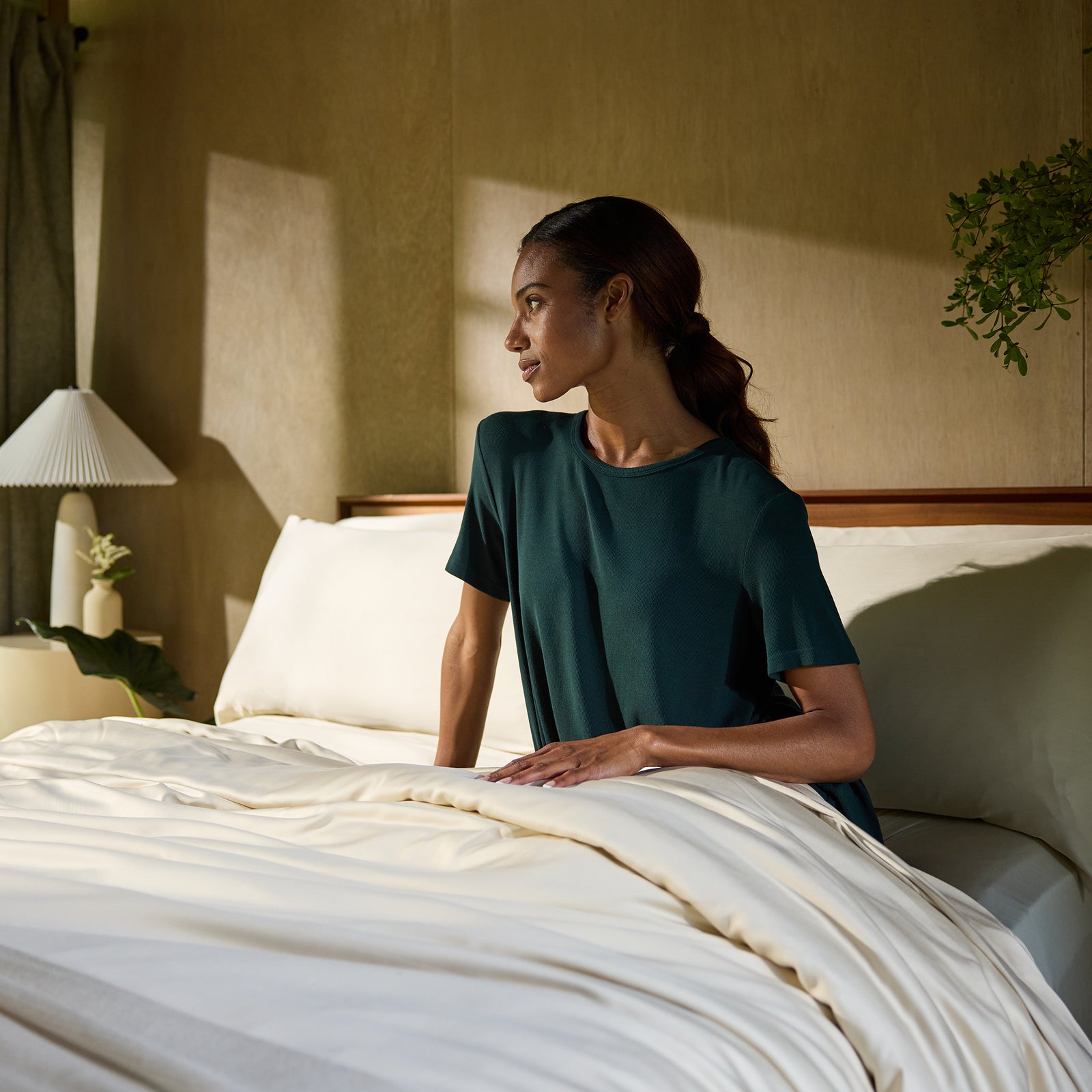 A person with long hair tied back sits up in bed, wearing a dark green short-sleeved shirt. They are looking out to the side with a relaxed expression. The bed is adorned with Cozy Earth's Bamboo Fitted Sheet, featuring light-colored fabric. The room is warmly lit with natural light, and a plant and lamp can be seen in the background. 