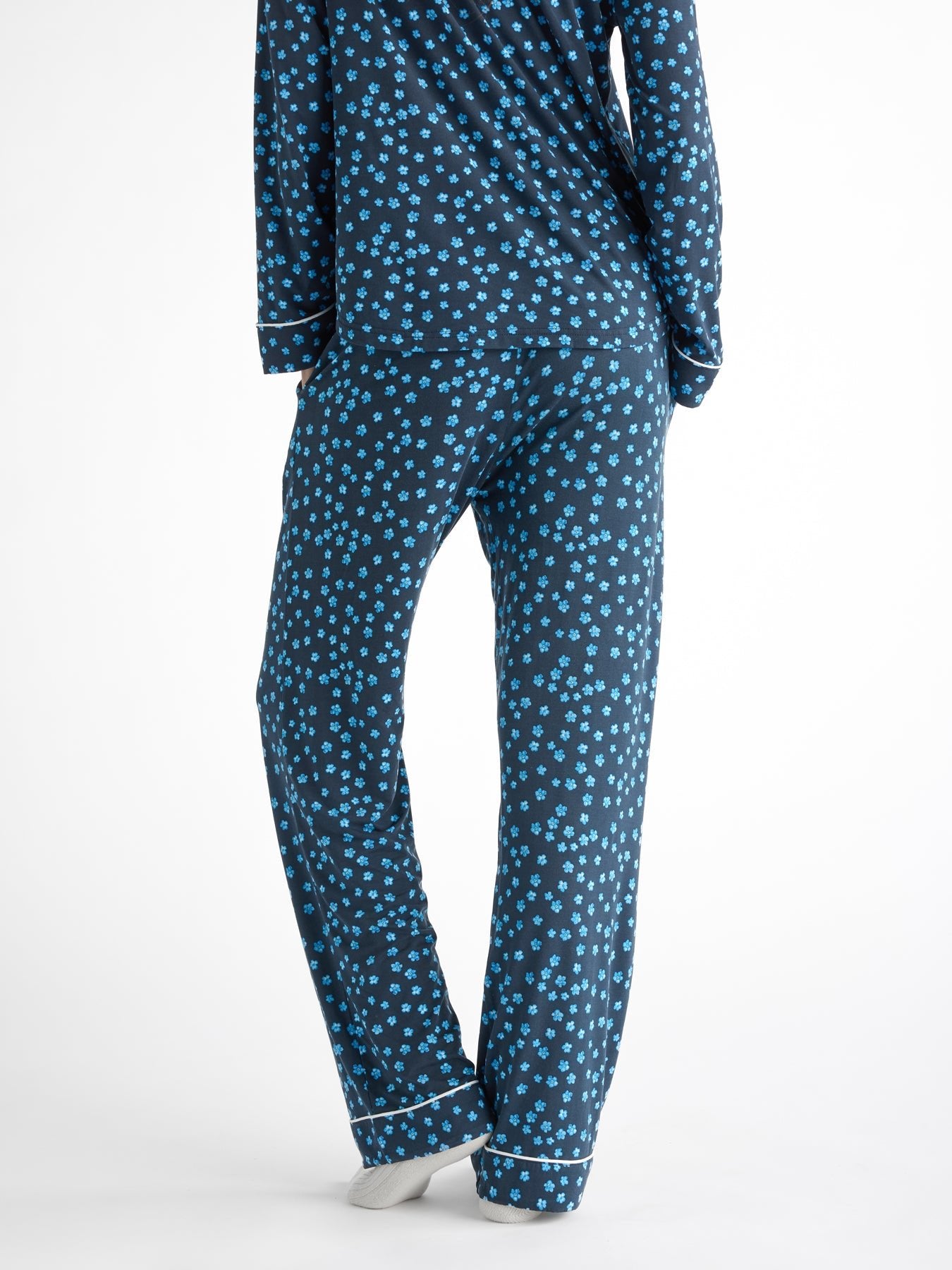 A person is wearing the Women's Bamboo Stretch-Knit Pajama Pant by Cozy Earth, featuring a dark blue background with a pattern of small blue flowers. The image captures their back view from the neck down, standing on a neutral-colored surface. 