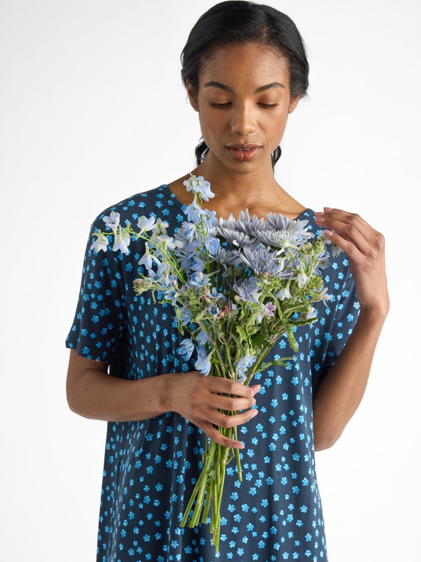 Wearing Cozy Earth's Women's Bamboo Stretch-Knit Sleep Dress, a person adorned in blue with delicate floral patterns gracefully holds a bouquet of blue and purple flowers, gazing serenely at them against a plain white background. 