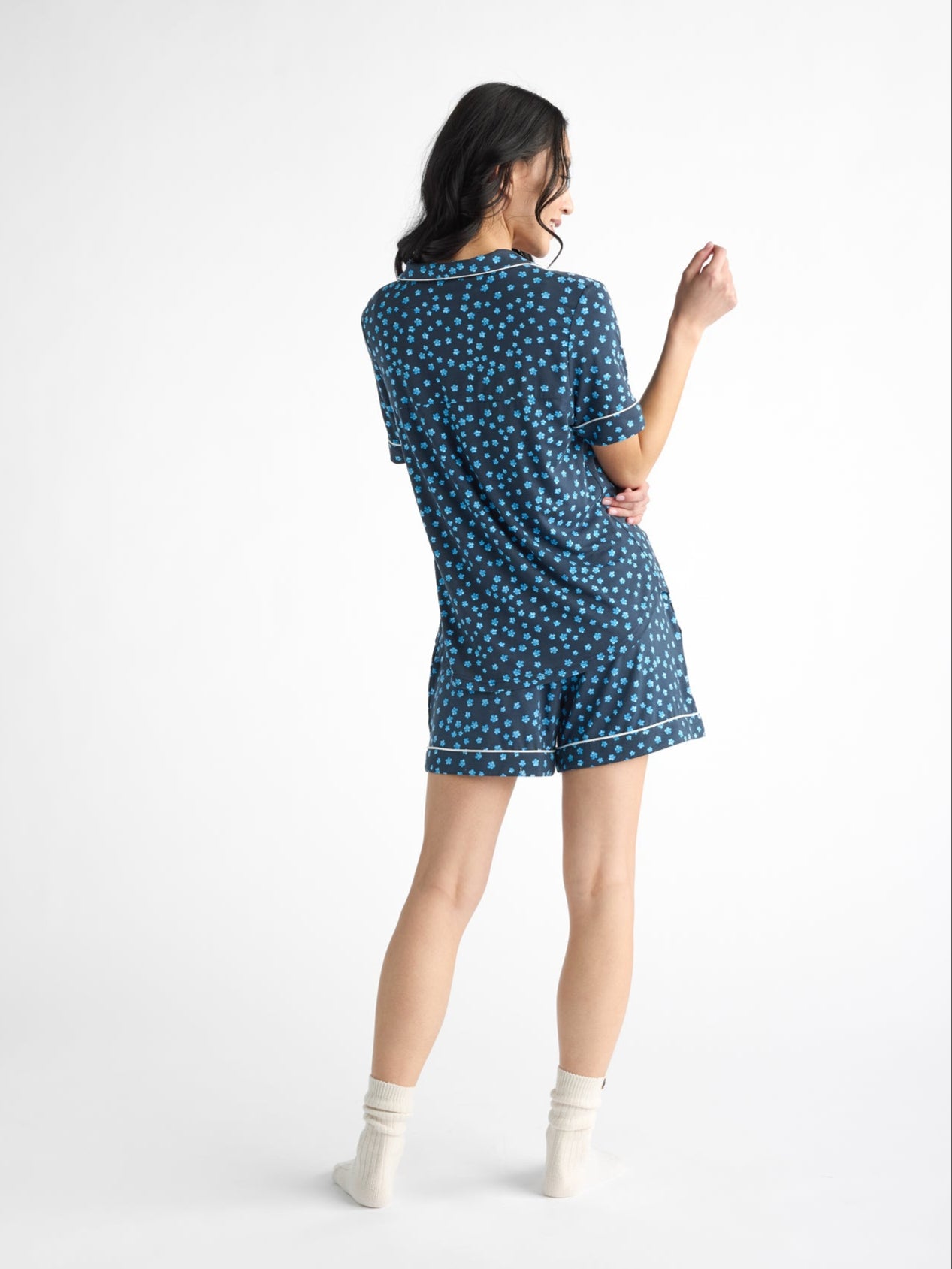 A person with shoulder-length dark hair stands barefoot, wearing white ankle socks and a blue Women's Bamboo Stretch-Knit Pajama Short from Cozy Earth, featuring a pattern of small, light blue dots. The background is plain white. 