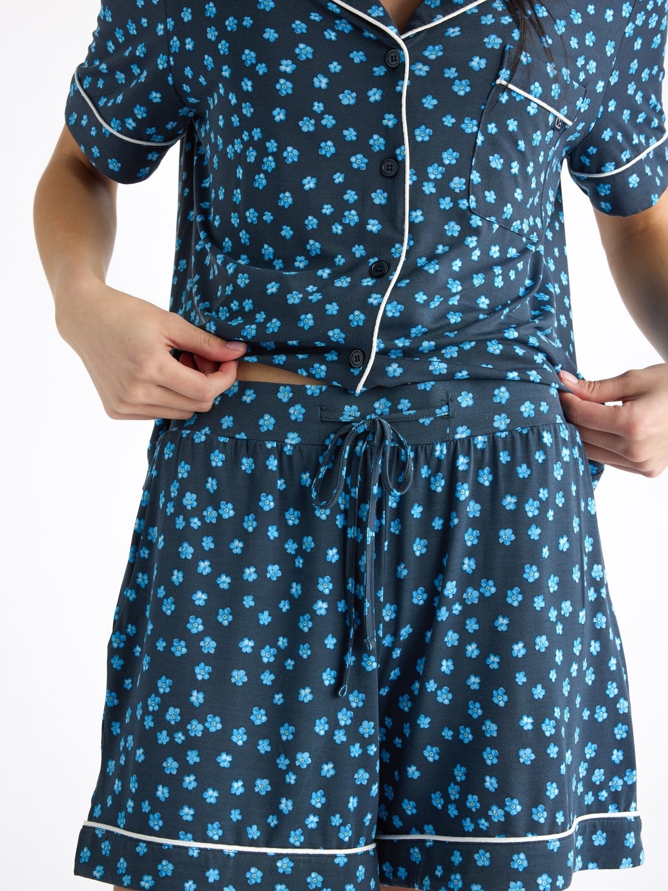 A person in the Women's Bamboo Stretch-Knit Short Sleeve Pajama Set by Cozy Earth, featuring a dark blue base with light blue floral patterns, adjusts the waistband of the elastic-waist shorts. The set includes a short-sleeved button-up top and is shown against a plain white background. 