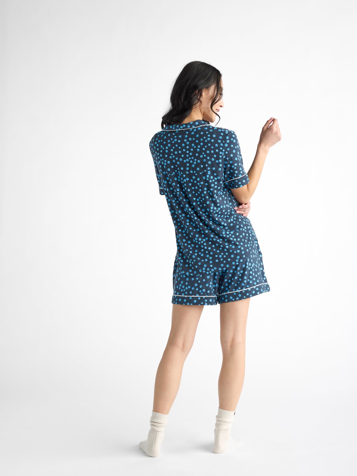 A woman with long dark hair, wearing a casual-fit Women's Bamboo Stretch-Knit Short Sleeve Pajama Top by HIDE and white socks, stands with her back turned against a plain white background. The blue pajama set features white polka dots. 