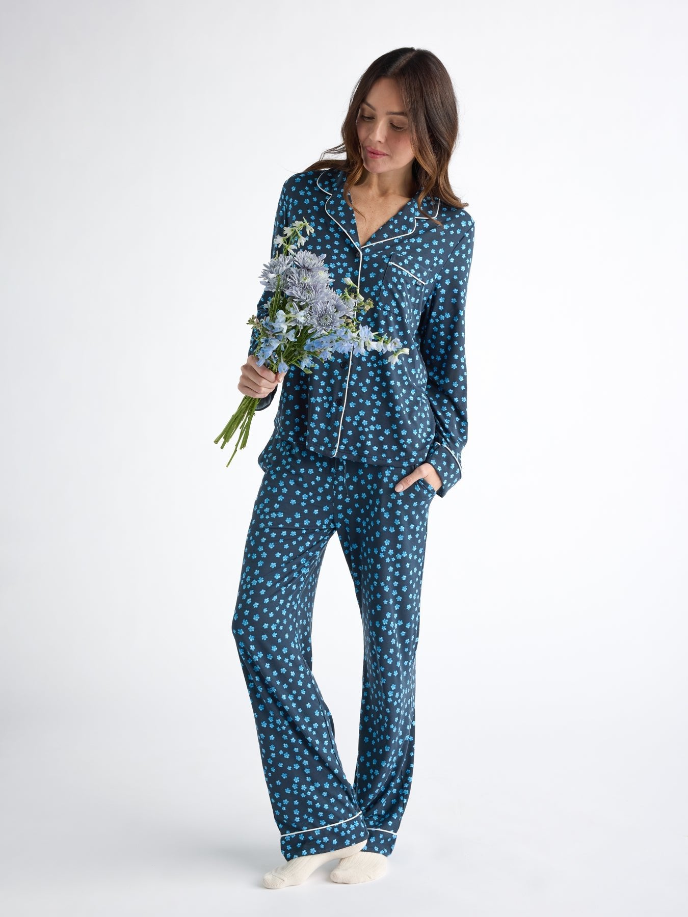 A woman in Cozy Earth's Women's Bamboo Stretch-Knit Long Sleeve Pajama Set, navy with white polka dots, holds flowers. Standing on a white background and looking down calmly, she rests her hand in her pocket and wears cream-colored socks. 