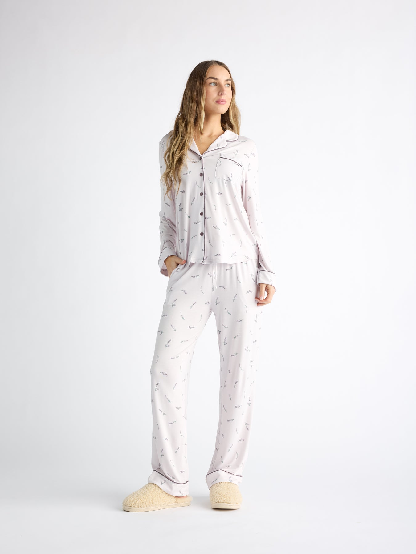 A person stands in Cozy Earth Women's Bamboo Stretch-Knit Pajama Pant, featuring small patterns and black piping and buttons. They wear fluffy slippers, all against a plain white background for a minimalist vibe. 