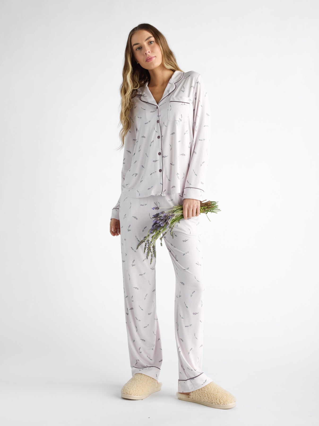 Against a white background, a person with long hair wears the light-colored Women's Bamboo Stretch-Knit Long Sleeve Pajama Set by Cozy Earth, holding purple flowers and cream slippers. 