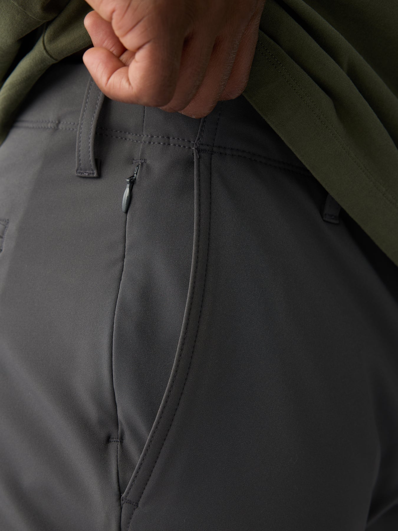 Close-up of a person wearing Cozy Earth's Men's Everywhere Pant in dark gray, highlighting the texture, stitching, and side zipper pocket detail while lightly gripping their green shirt. 