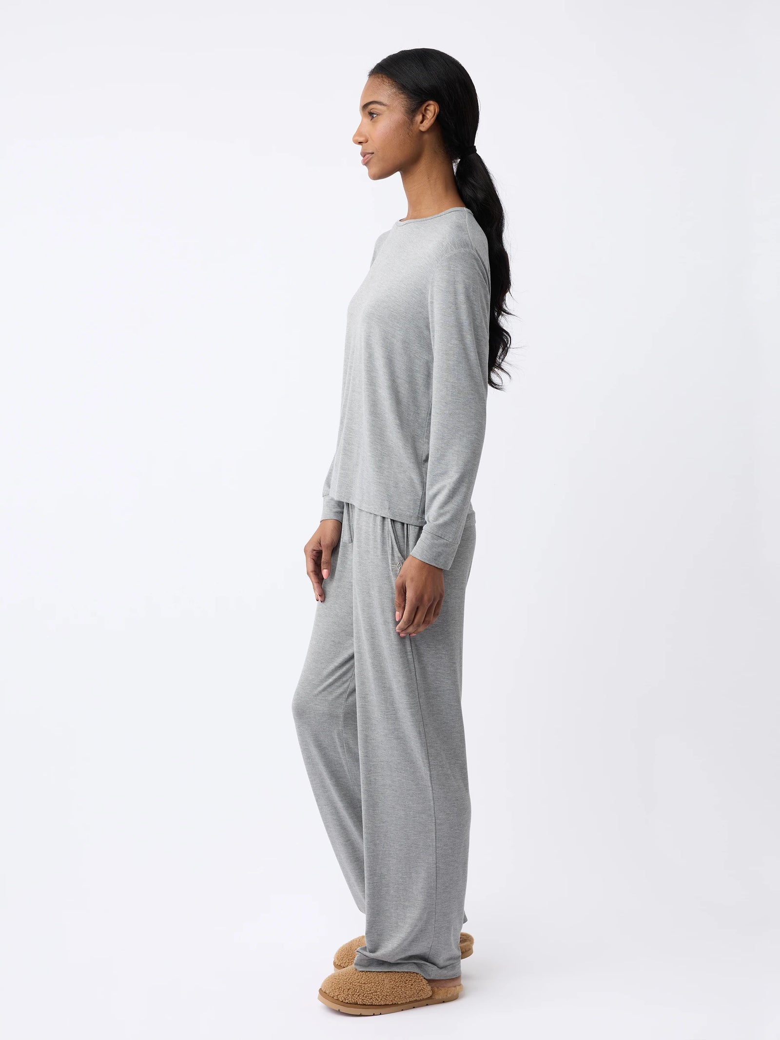 A woman in a gray Women's Stretch Knit Bamboo Long Sleeve Lounge Tee by Cozy Earth and tan slippers stands sideways against a white background, with long hair tied back in a ponytail. 