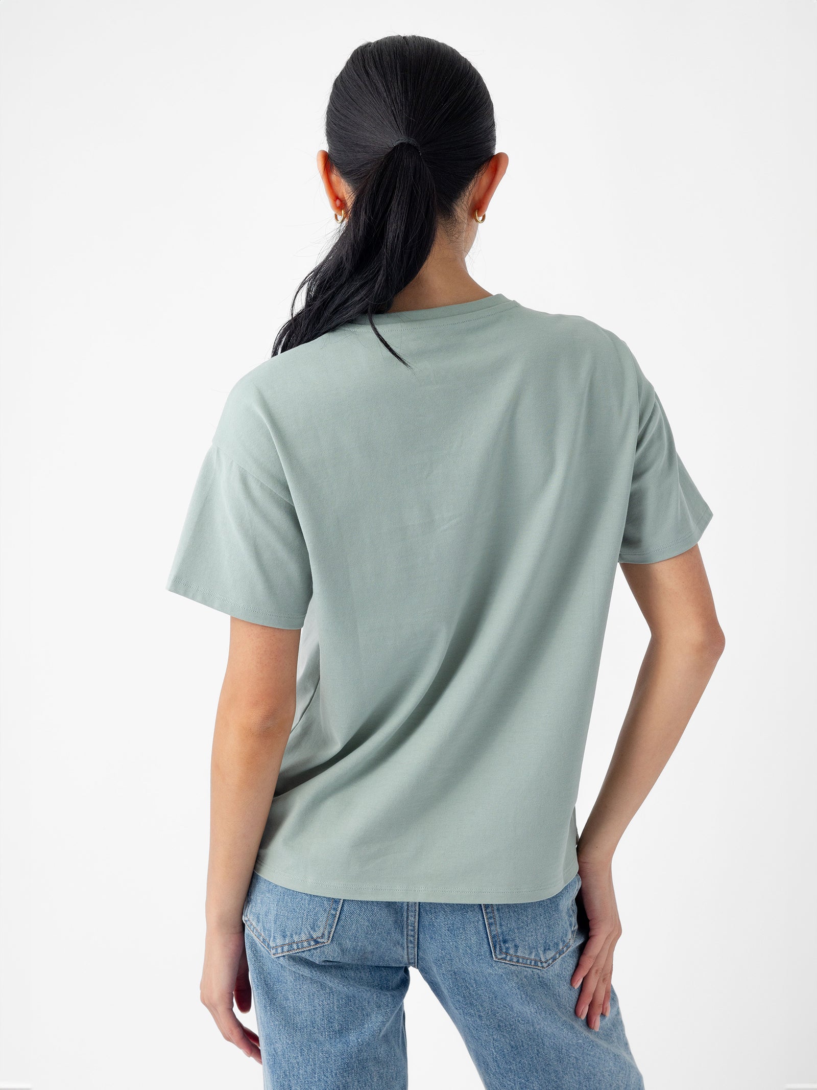 A person with long black hair in a ponytail is standing with their back to the camera, wearing a Cozy Earth Women's All Day Tee in light green and blue jeans. The background is plain white. 