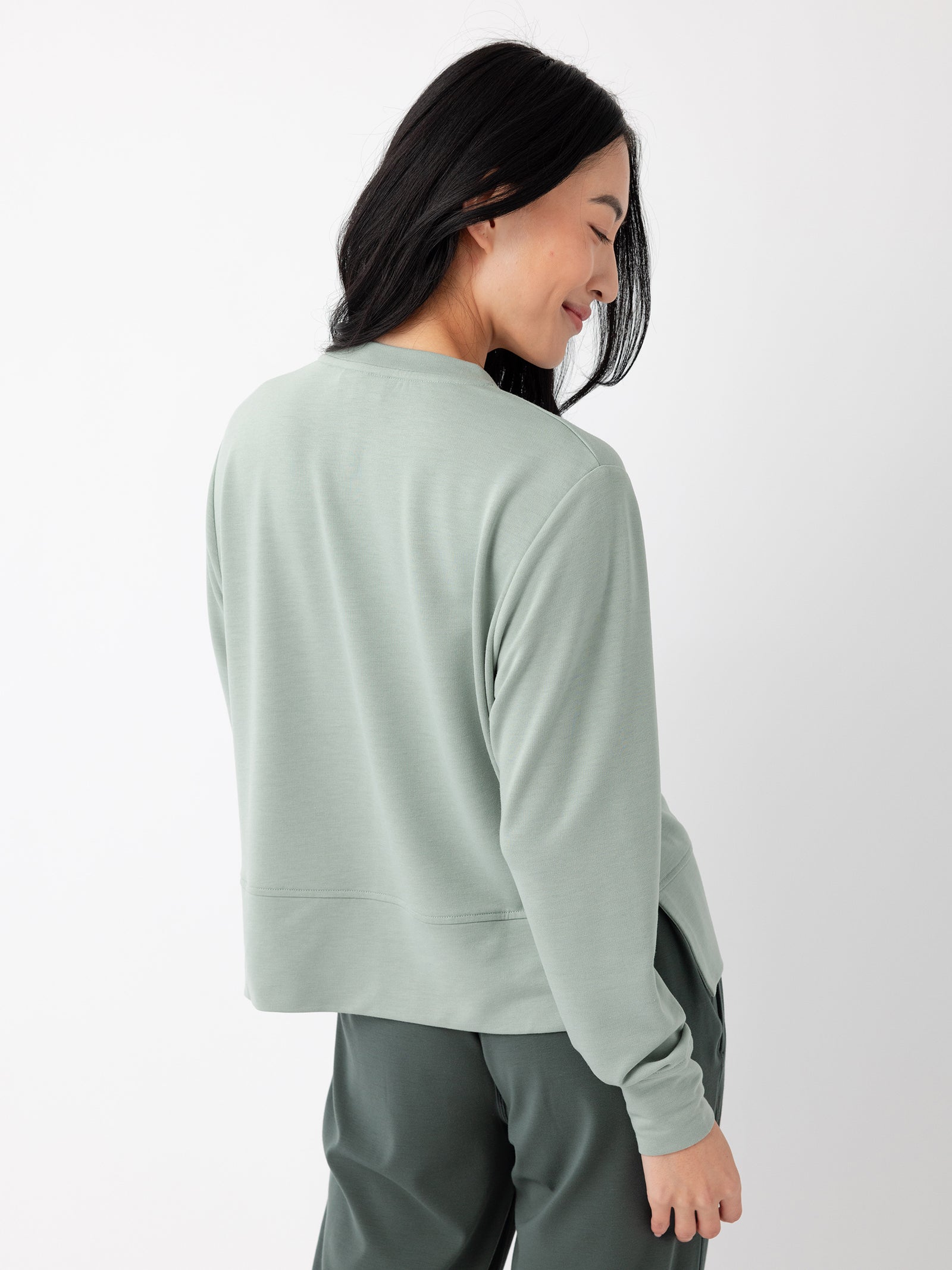 A woman with long, dark hair is standing and smiling, looking over her shoulder. She is wearing Cozy Earth's Women's StudioLite Crewneck in a loose, light green long-sleeved top paired with dark green pants. The background is plain white. 