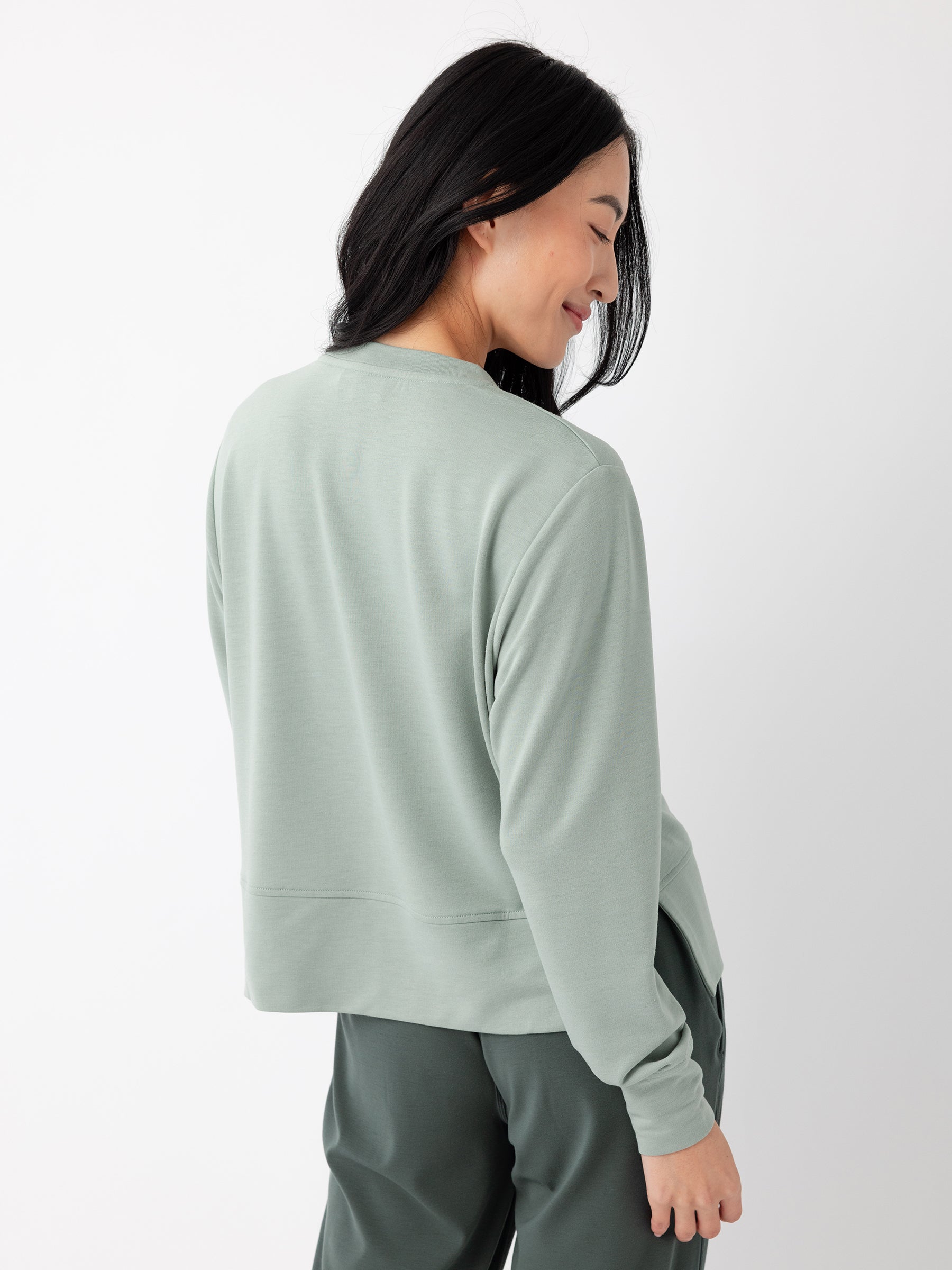 A woman with long, dark hair is standing and smiling, looking over her shoulder. She is wearing Cozy Earth's Women's StudioLite Crewneck in a loose, light green long-sleeved top paired with dark green pants. The background is plain white. |Color:Haze