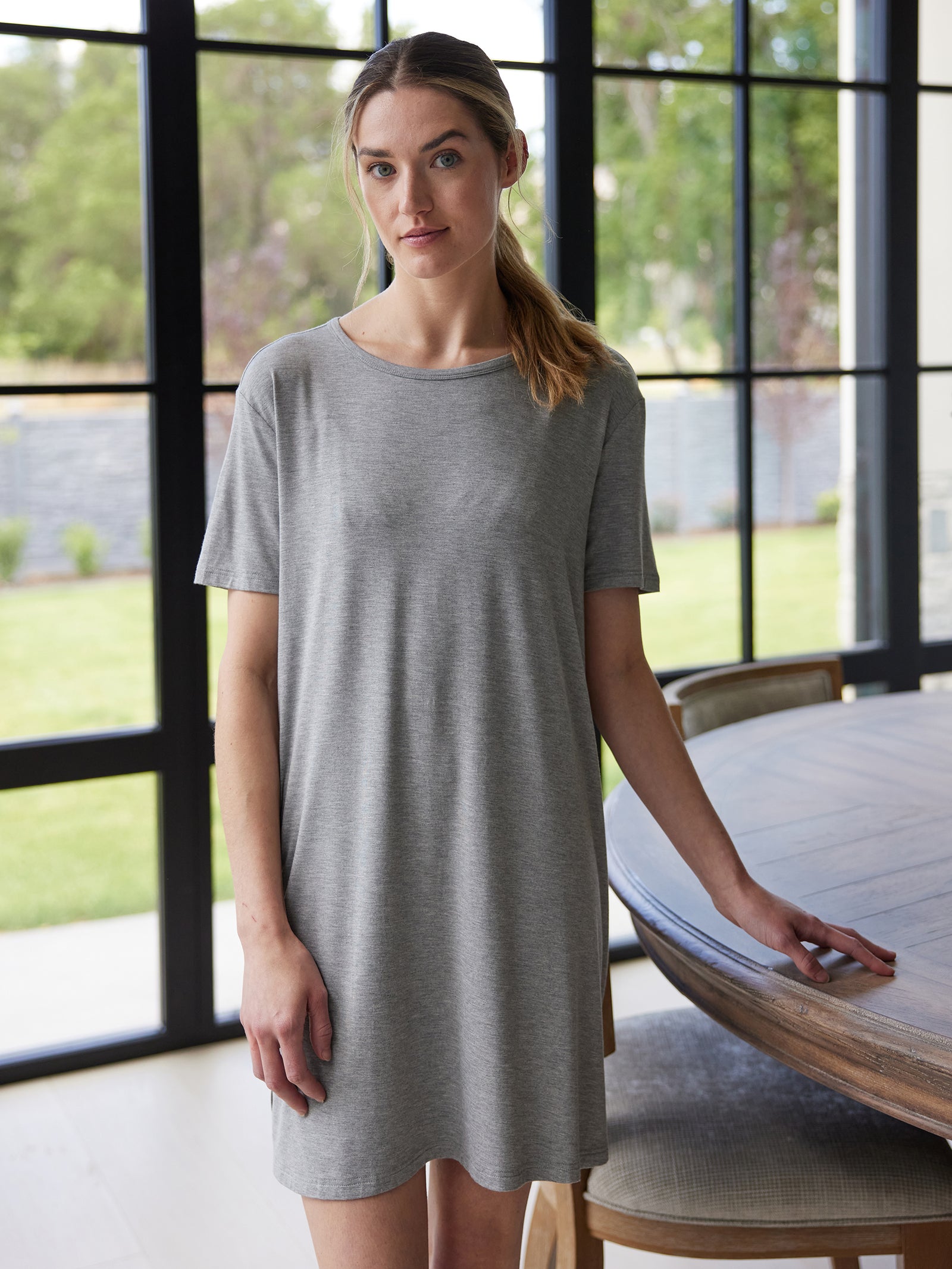 A woman in a gray Cozy Earth Women's Bamboo Stretch Knit Sleep Dress stands indoors near a round wooden table. Large windows in the background reveal a grassy outdoor area. She has a neutral expression and her light brown hair is pulled back in a loose ponytail. 