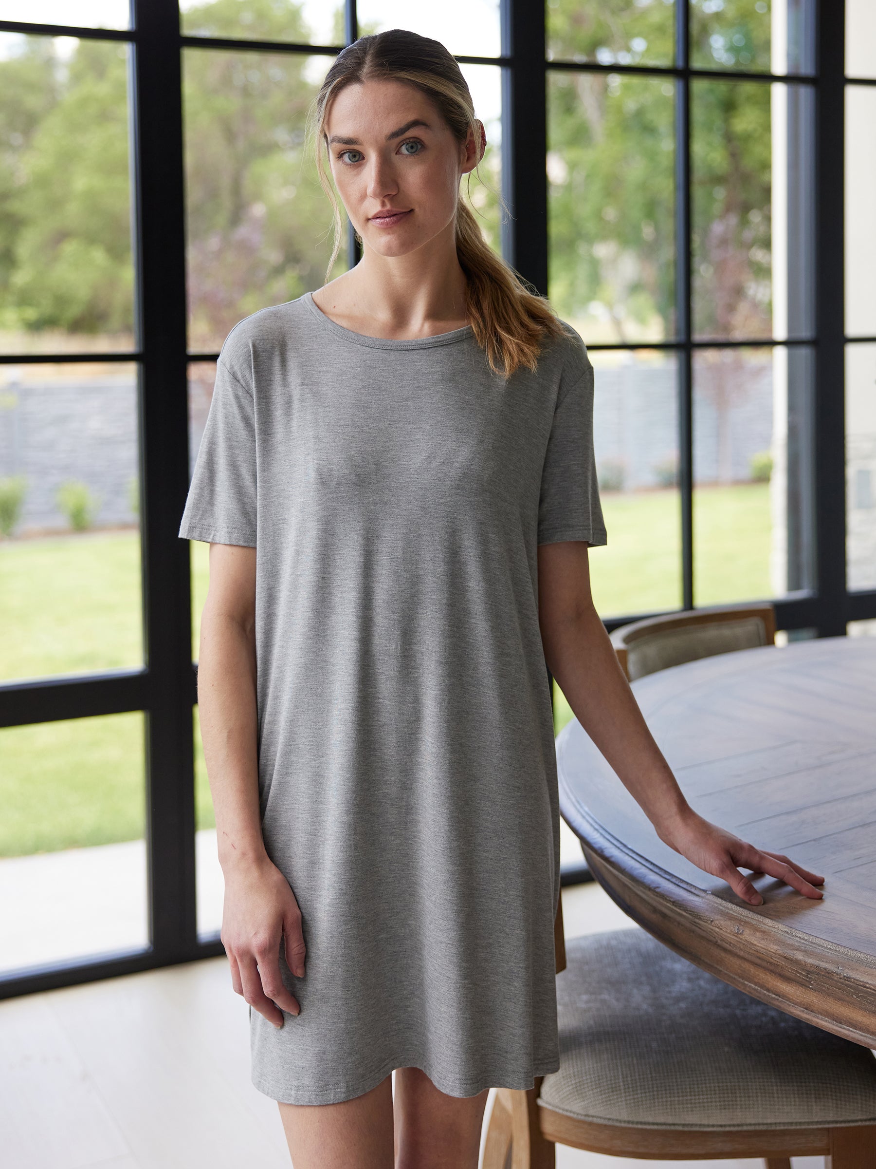 A woman in a gray Cozy Earth Women's Bamboo Stretch Knit Sleep Dress stands indoors near a round wooden table. Large windows in the background reveal a grassy outdoor area. She has a neutral expression and her light brown hair is pulled back in a loose ponytail. |Color:Heather Grey