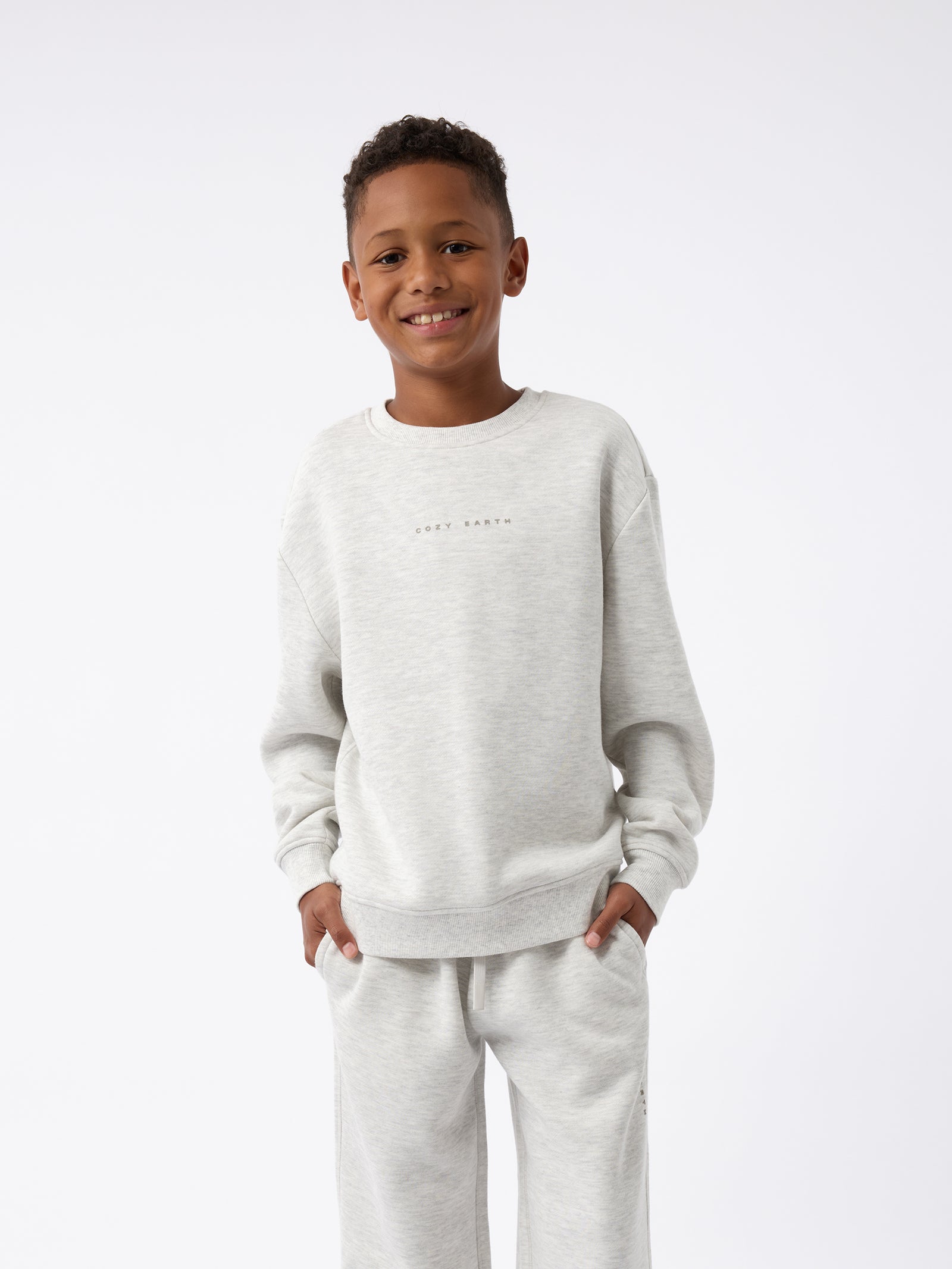 A smiling boy stands against a plain background, wearing the Cozy Earth Kid's CityScape Crewneck in light gray, paired with matching sweatpants. With his hands in his pockets, he exudes a casual, relaxed vibe. 