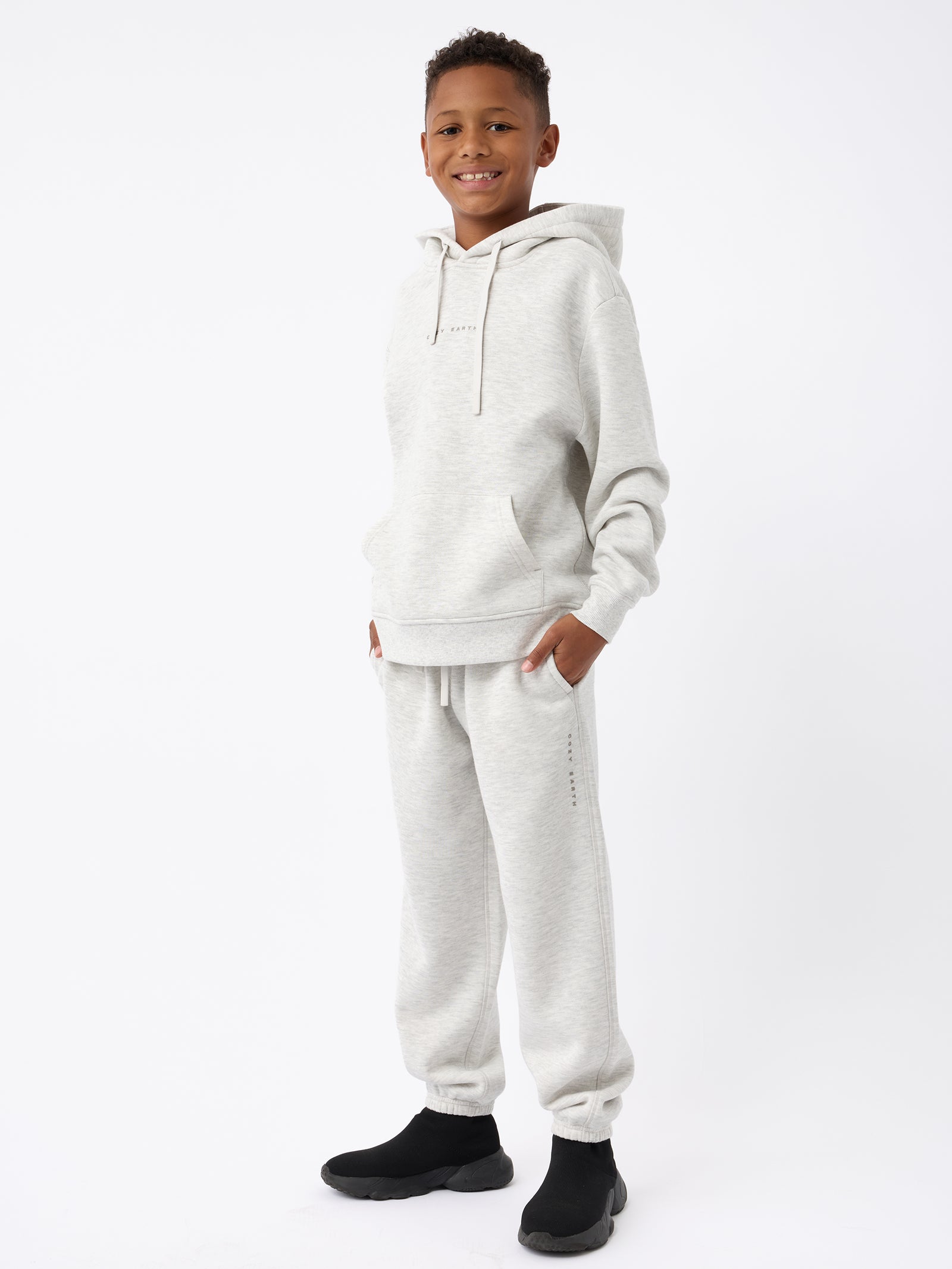 A smiling boy wearing a Cozy Earth Kid's CityScape Hoodie and matching sweatpants stands against a white background. He has his hands in the hoodie pockets and is wearing black shoes. His light gray hoodie features a small printed logo. 