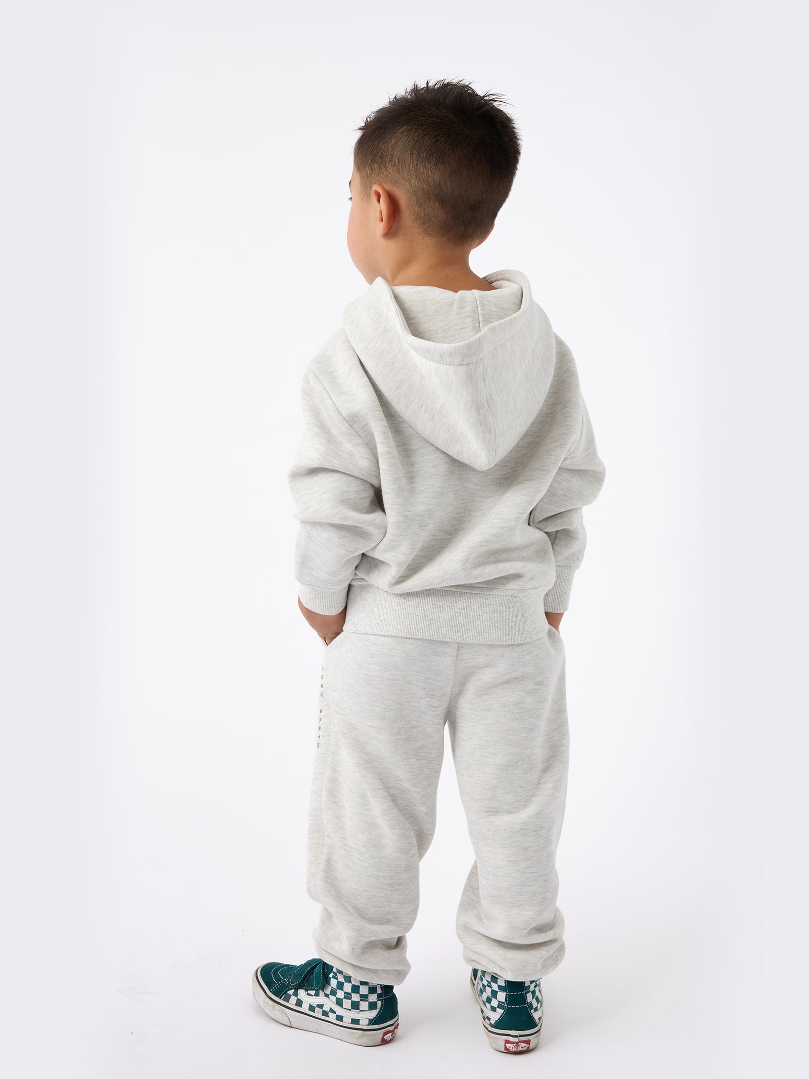 A young child stands facing away, dressed in Cozy Earth's Kid's CityScape Sweatpant and a light gray hoodie. They have short dark hair and are sporting green and white checkered sneakers against a plain white backdrop. 