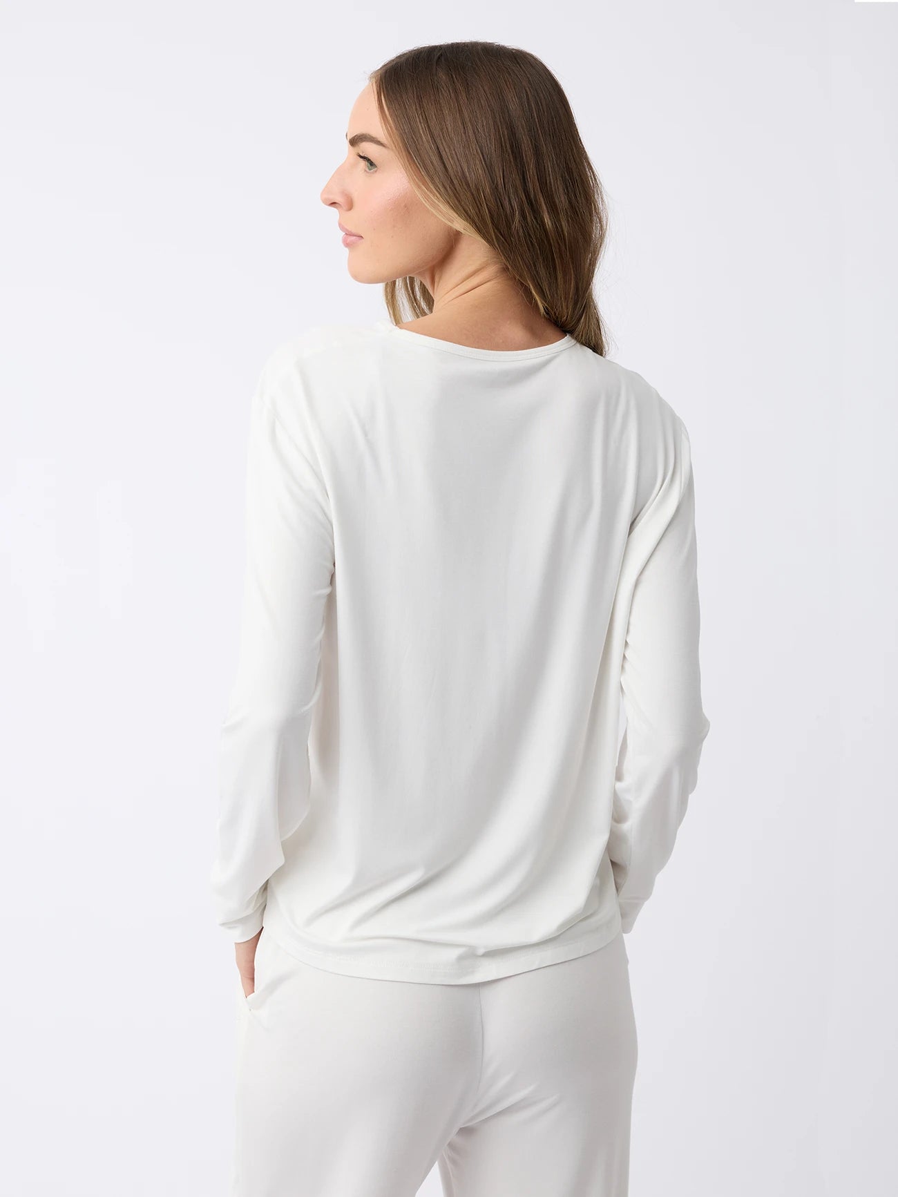 A woman with long hair stands facing away in a plain, light-colored setting, wearing the Cozy Earth Women's Stretch Knit Bamboo Long Sleeve Lounge Tee paired with matching pants. 