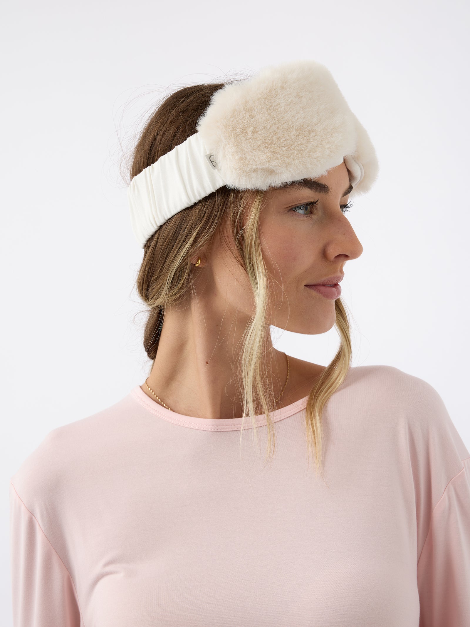 A woman wears the Cozy Earth Faux Fur Sleep Mask in Ivory. The mask is positioned on her forehead, above her eyes. 