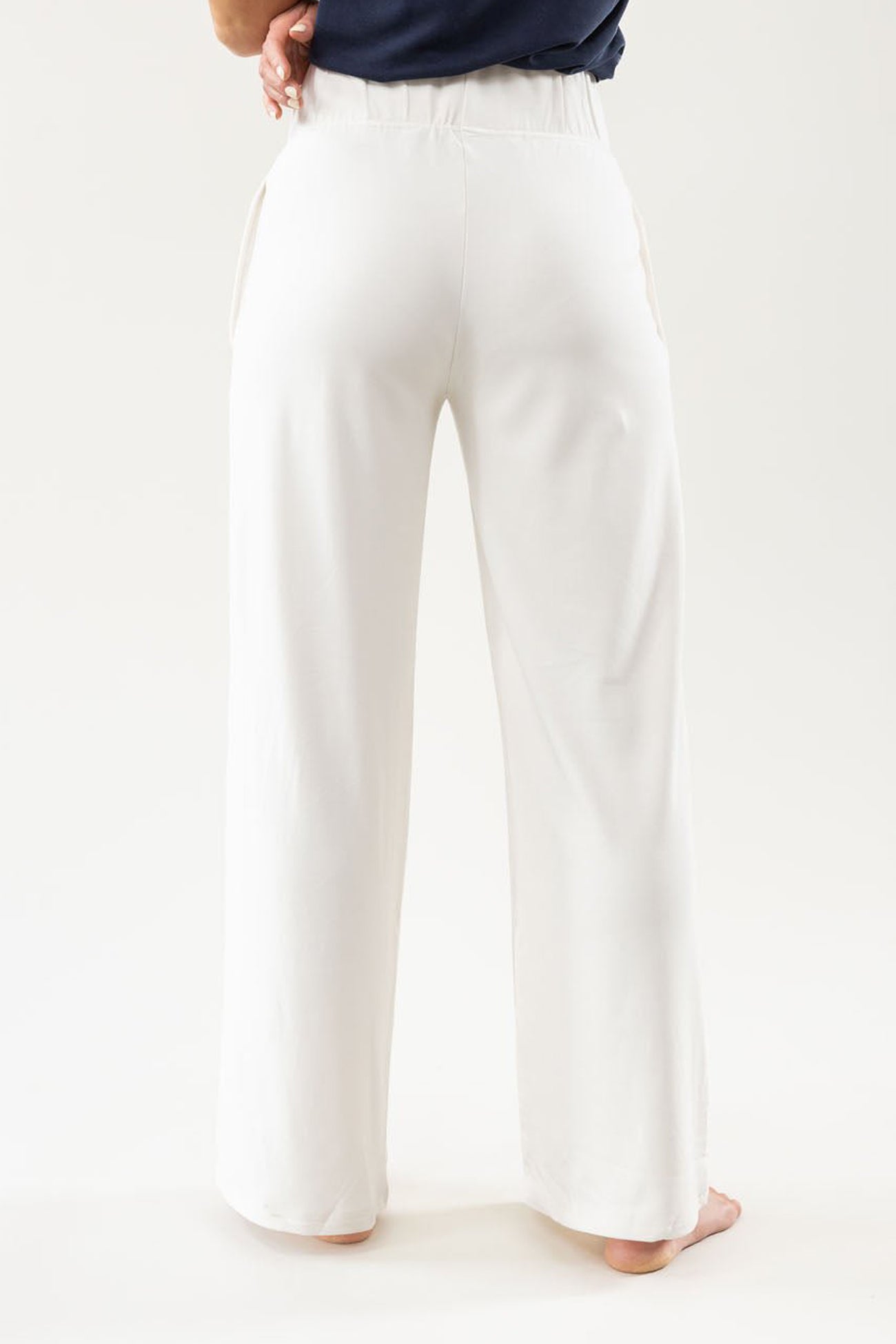 A person is wearing white Cozy Earth's Women's Ultra-Soft Bamboo Wide Leg Pull On Pant, featuring an elastic waistband and pockets. The view is from the back, barefoot on a light-colored floor. 