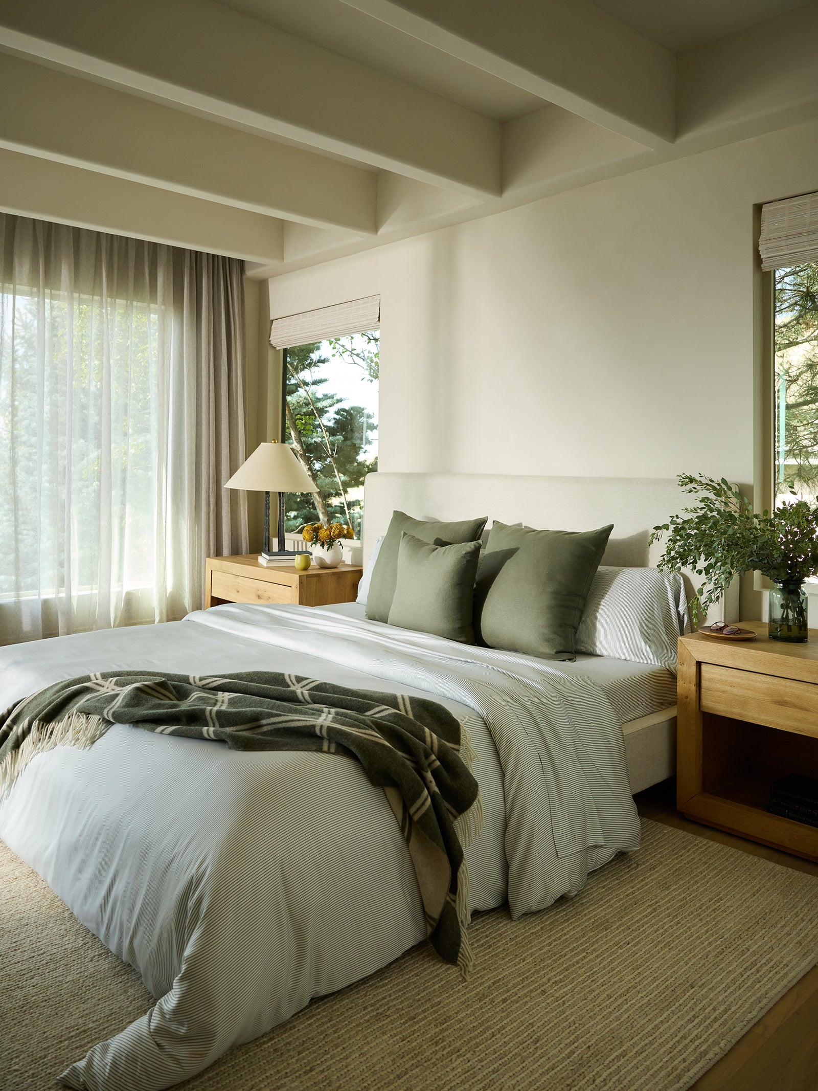 The tranquil bedroom showcases a neatly made bed dressed in white linens, accented by green pillows made with Bamboo Pillowcases from Cozy Earth. A matching green throw is casually draped over one corner of the bed. The warm atmosphere is complemented by wooden nightstands adorned with lamps and a plant. Ample natural light floods the room through large windows, creating an inviting ambiance. 