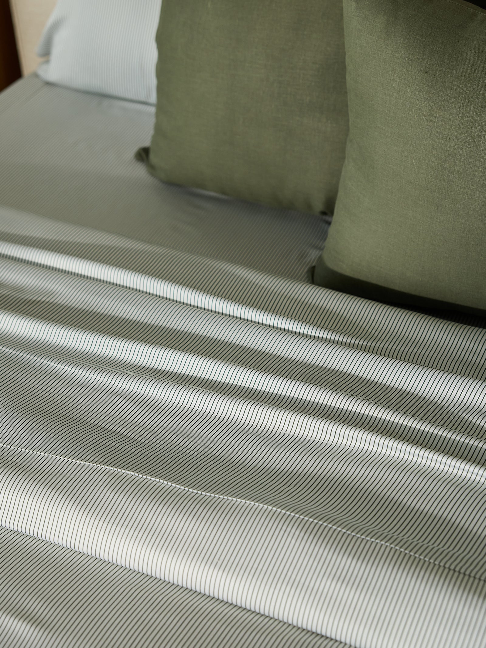 A neatly made bed features a Bamboo Fitted Sheet from Cozy Earth in gray and white stripes. Two olive green pillows and a light blue pillow are arranged at the head of the bed, creating a calm and inviting ambiance. 