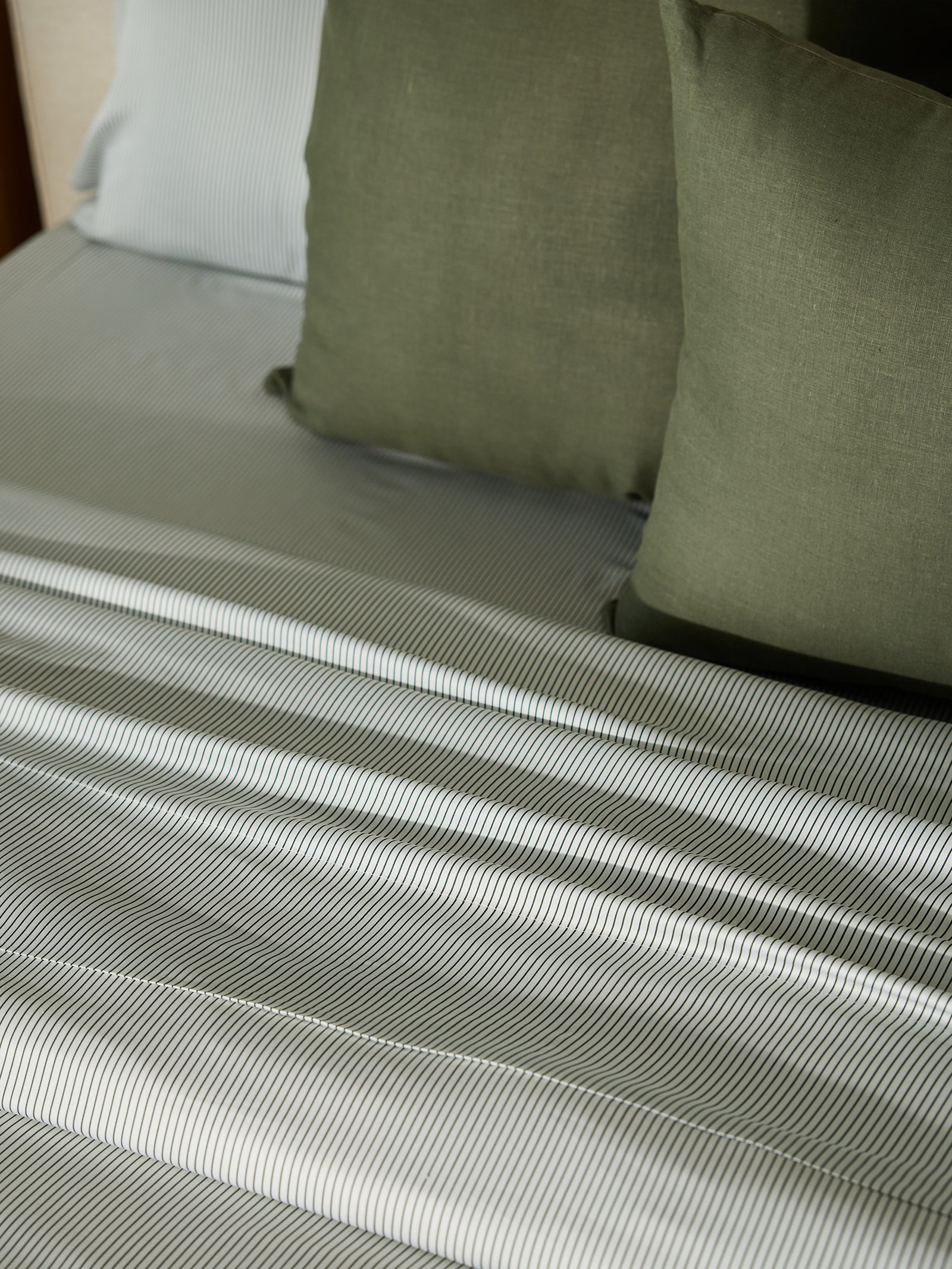 A close-up showcases a neatly made bed featuring Cozy Earth's Bamboo Flat Sheet with its striped linens in shades of gray, complemented by three olive green pillows, creating an atmosphere of comfort and modern style. 