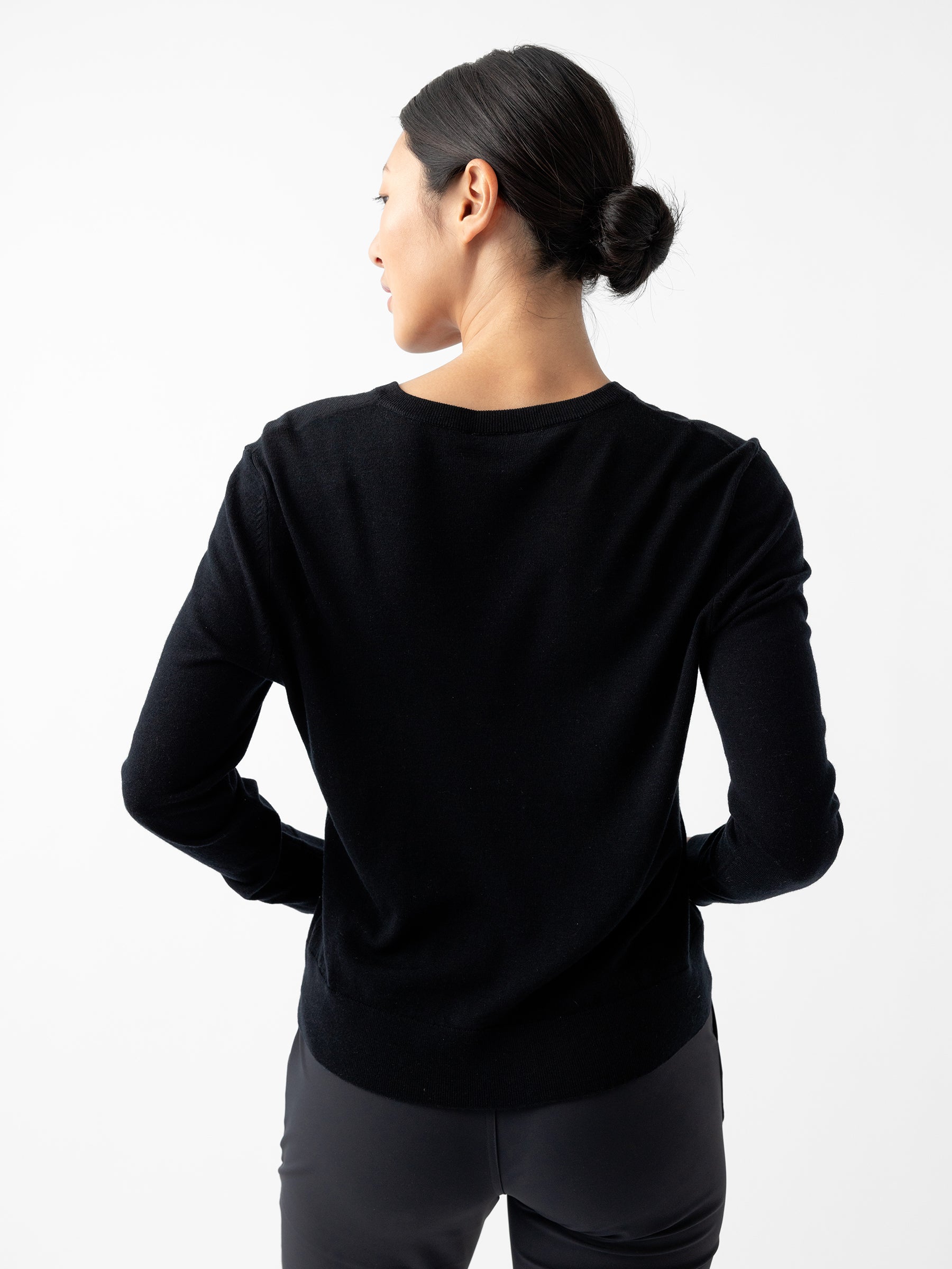 A person with dark hair tied in a low bun is wearing a black Women's AirKnit V-Neck Sweater from Cozy Earth and dark pants. They are standing facing away from the camera against a plain white background, with their hands placed on their hips. |Color:Jet Black