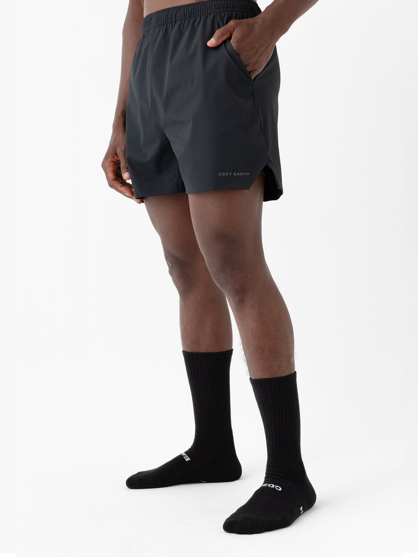 A person is wearing Cozy Earth's Men's Performance Sleep Shorts and black socks, standing against a plain white background. The shorts have the words "GREY EARTH" printed on the left leg. The person has their right hand in their pocket. Only the lower part of the body is visible. 