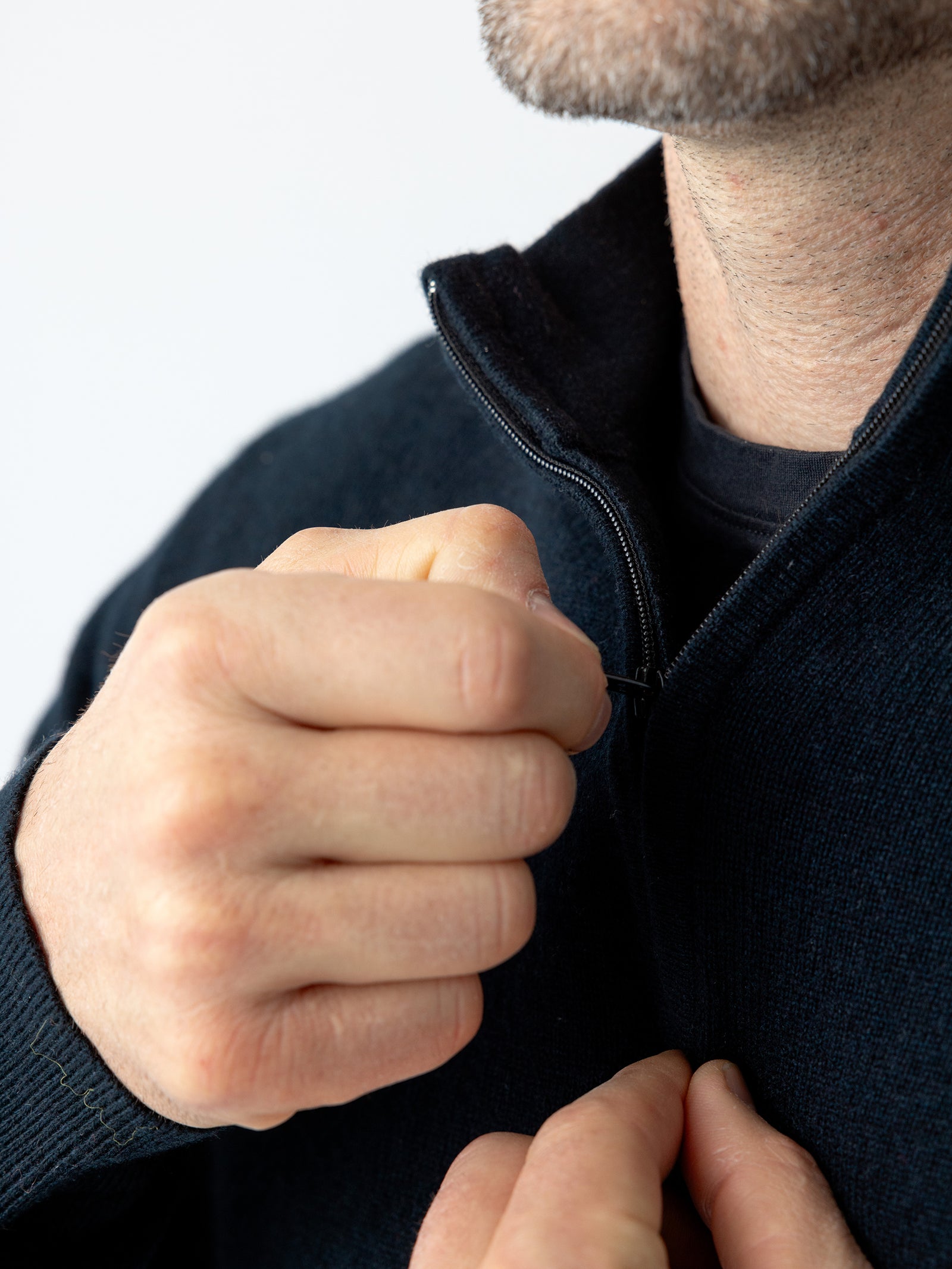 Close-up of a person zipping up a Cozy Earth Men's Quarter Zip Sweater. The person's hands are holding the zipper, and part of their face with a beard and mustache is visible. The background is plain and blurred. 