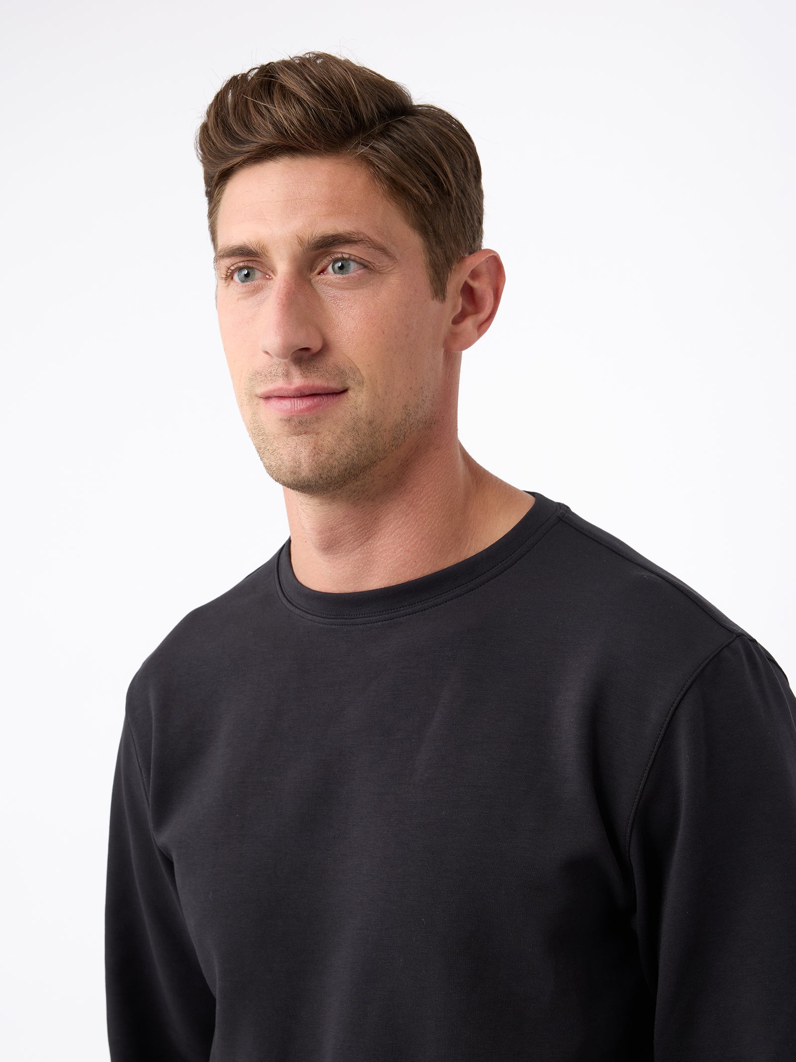 A man with short, brown hair and blue eyes is wearing a Cozy Earth Men's StretchTech Crewneck in black, gazing slightly to the left. The plain white background adds a clean and minimalistic feel to the image. 