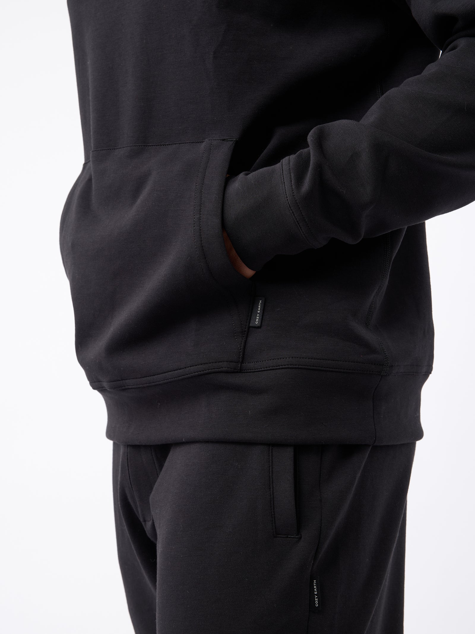 A person wearing a Cozy Earth Men's StretchTech Hoodie in black has one hand in the front pocket. The person is also wearing matching black pants. The background is plain white, showing only the torso and upper legs. 