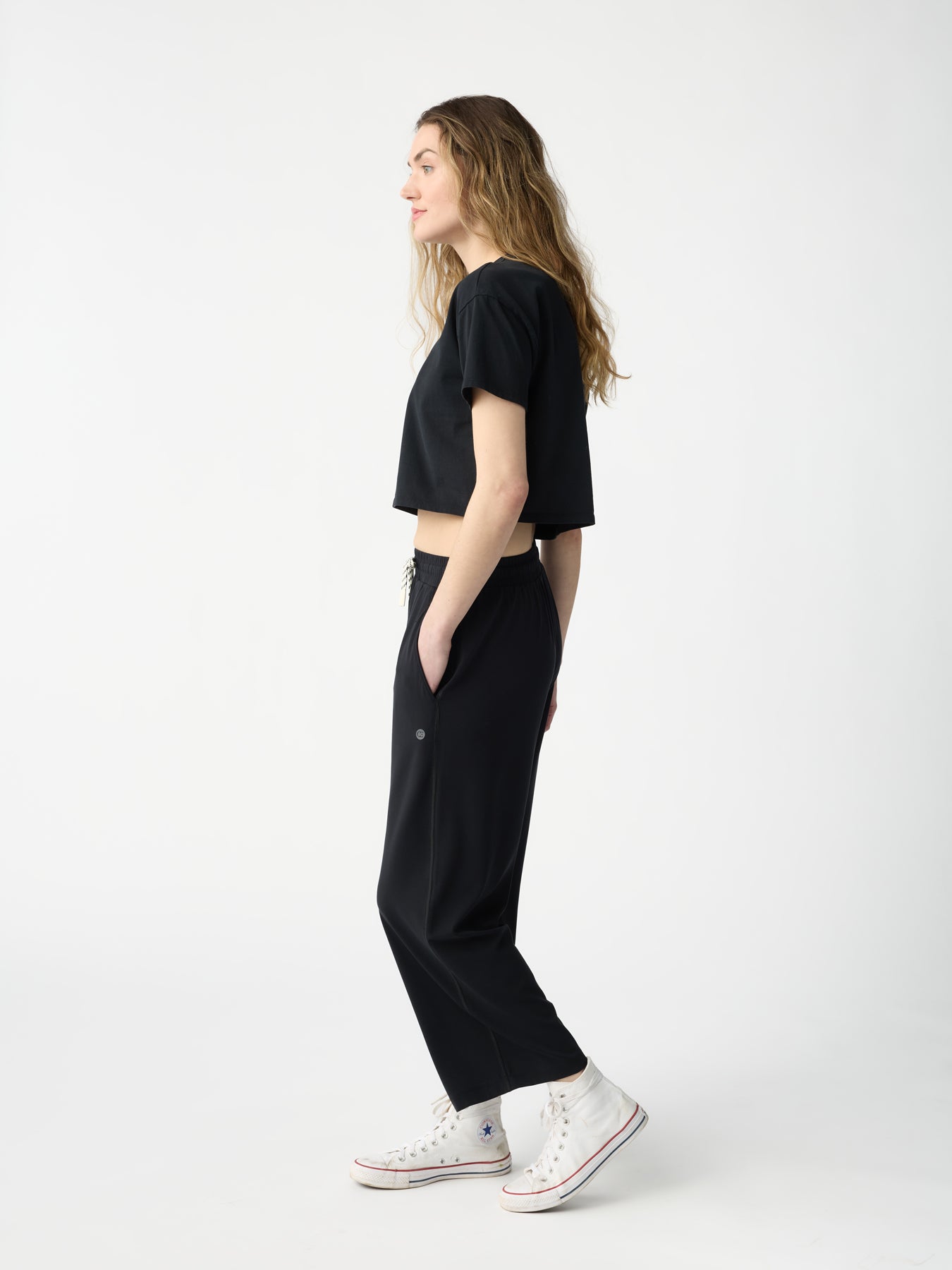 A person with long hair stands in profile against a plain, light background wearing a black cropped T-shirt, Women's Studio Cropped Wide Leg Pant by Cozy Earth, and white high-top sneakers. Their hands rest in their pockets. |Color:Jet Black