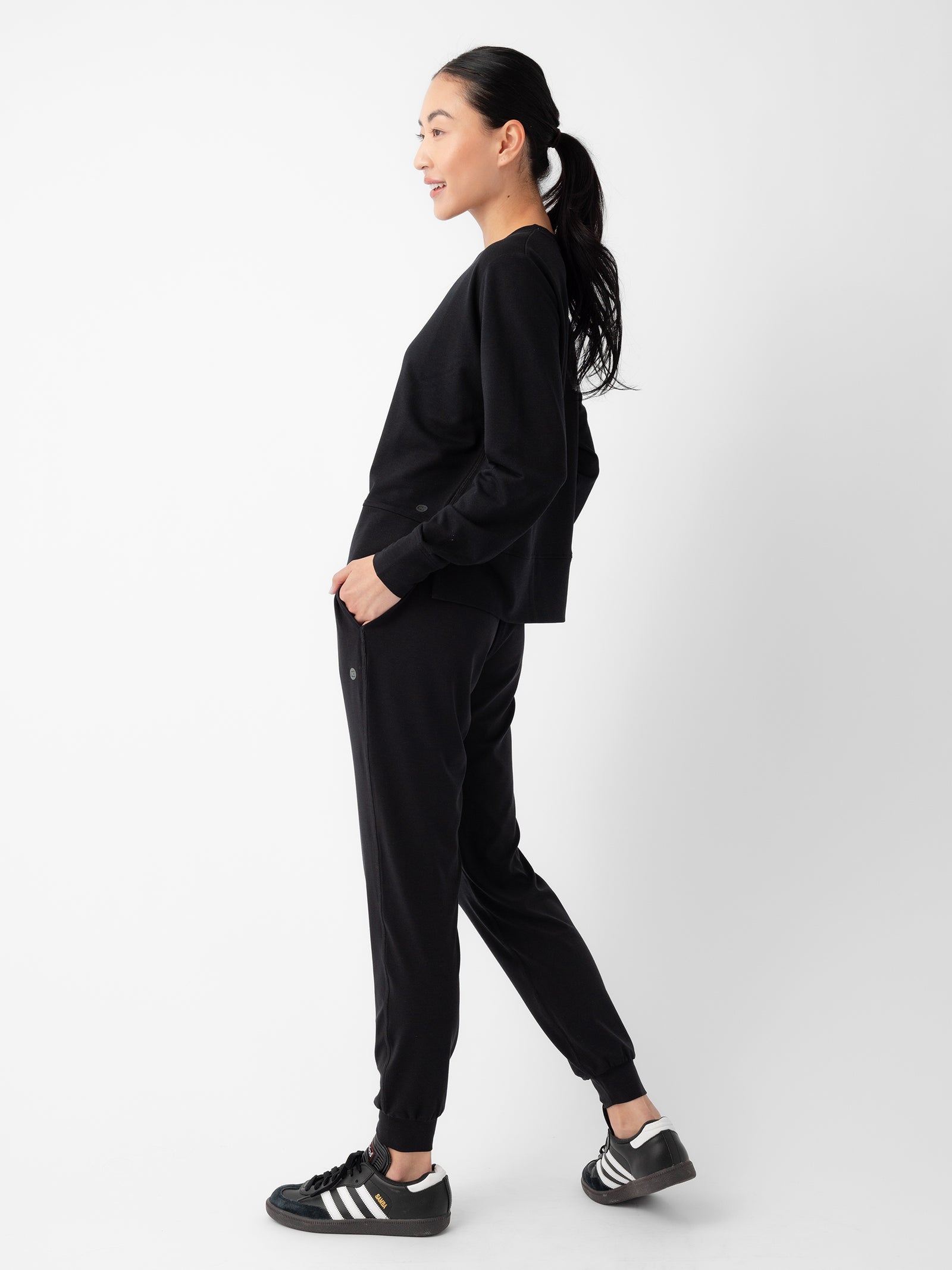 A person with long dark hair tied back in a ponytail is standing sideways against a plain white background. They are wearing the Women's StudioLite Crewneck by Cozy Earth, black jogger pants, and black Adidas sneakers with white stripes. 