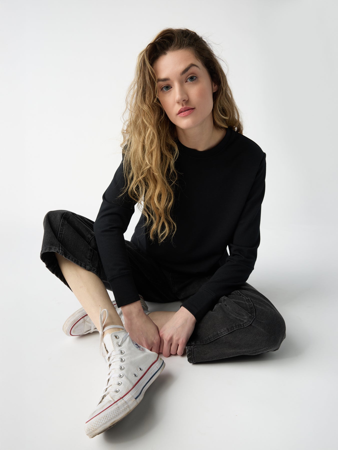 A person with long, wavy hair sits cross-legged on the ground, wearing a black Cozy Earth Women's StudioLite Crewneck, dark jeans, and white high-top sneakers. The background is plain white. 