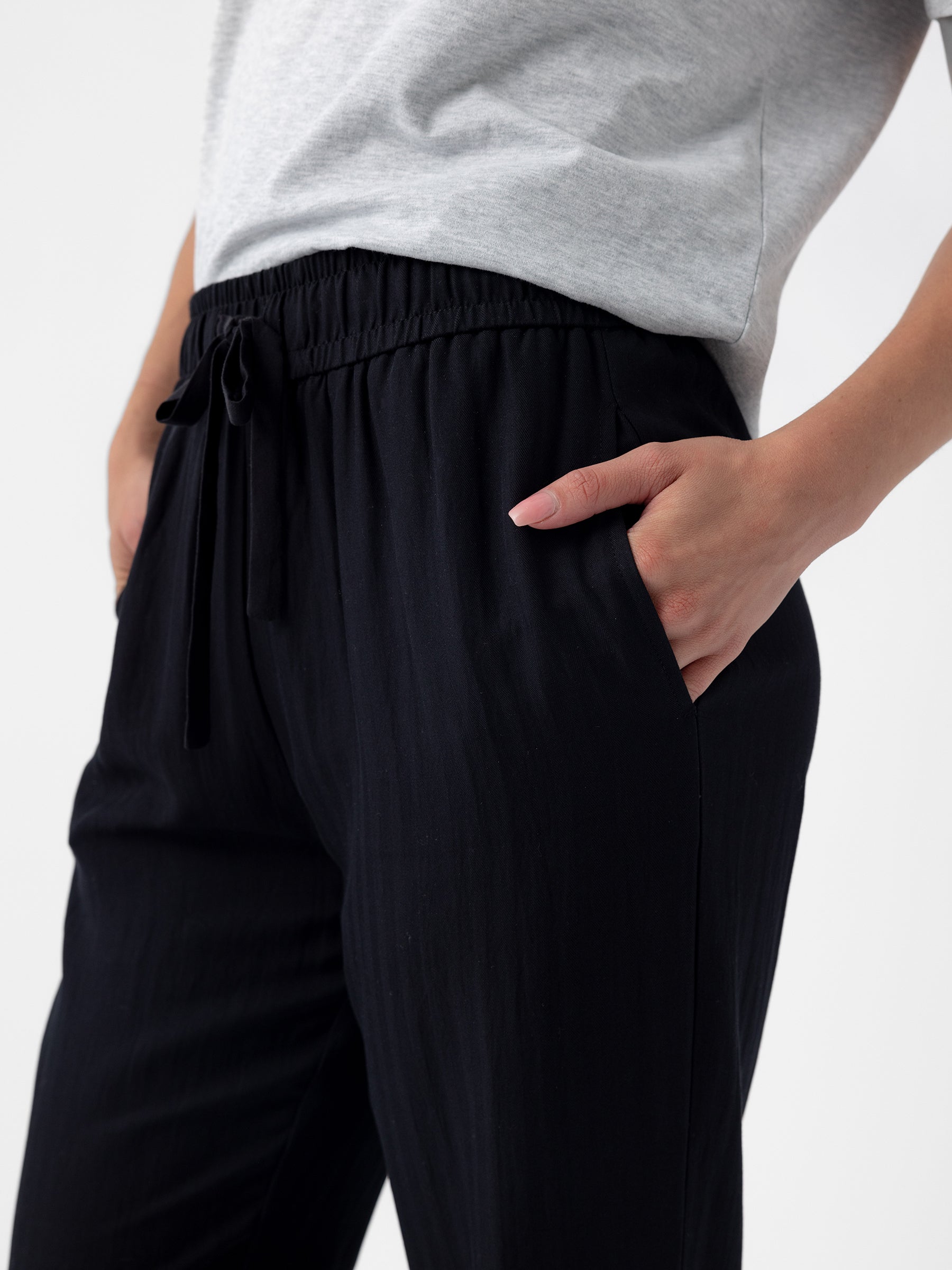 Close-up of a person wearing a casual outfit. They have on a gray t-shirt and Cozy Earth's Women's Sunset Cropped Pant in black, with their right hand inside the pants pocket. The background is plain and white. |Color:Jet Black